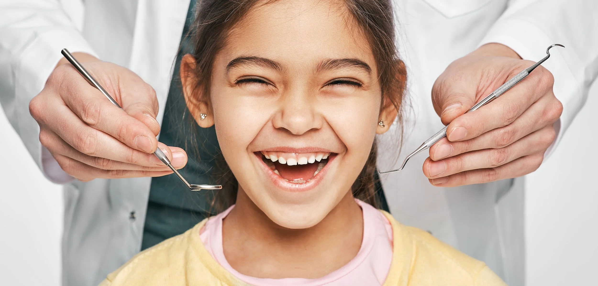 A young girl smiling at the dentist's chair while a dentist examines her teeth with dental tools.