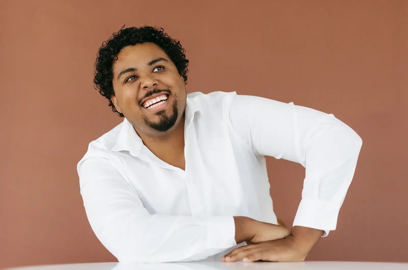 A man with curly black hair and a goatee, wearing a white shirt, smiling and looking to the side against a plain burnt orange background.