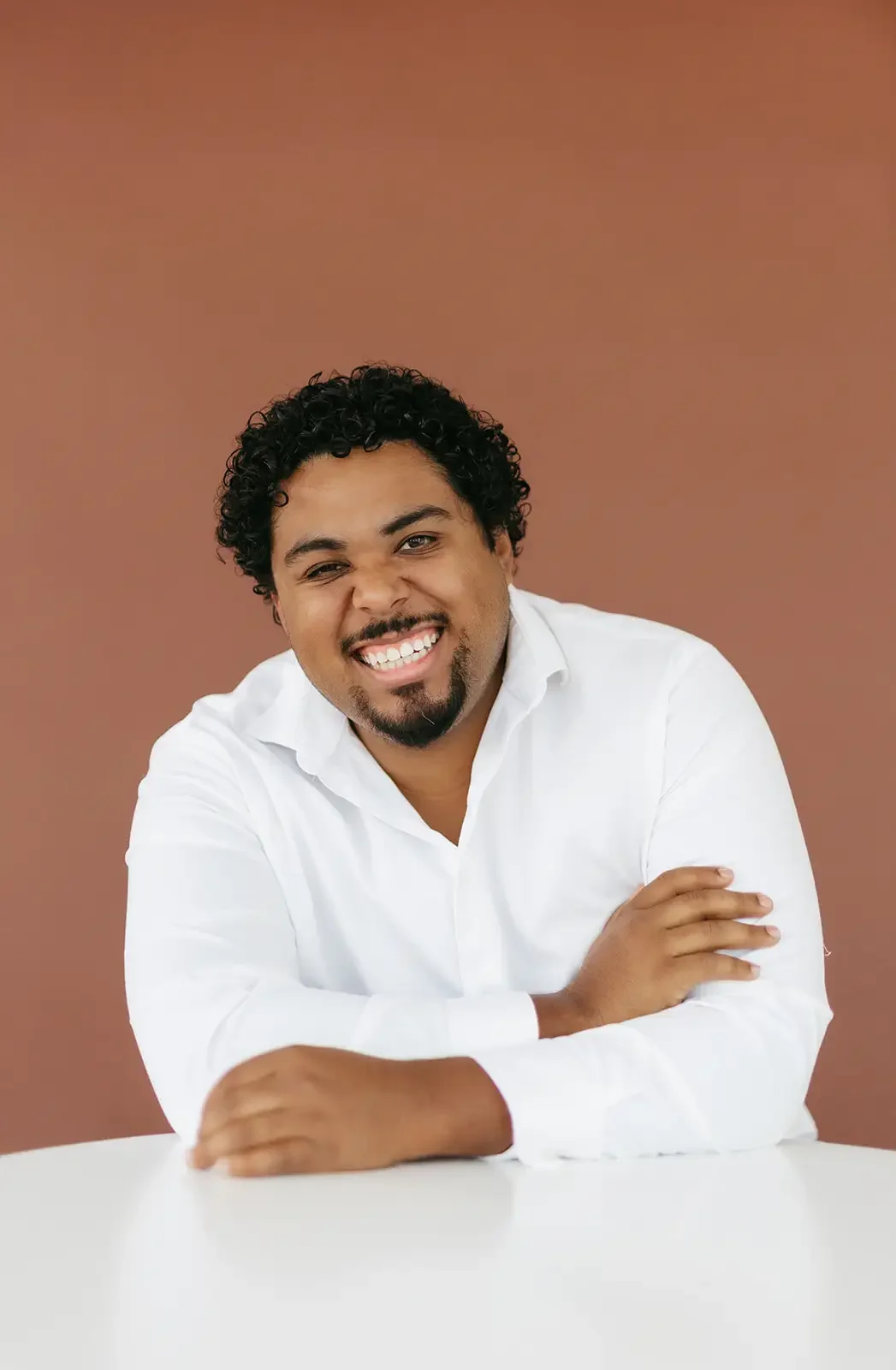 A tan man with curly hair and a goatee, wearing a white shirt, smiling and sitting at a table against a burnt orange background.