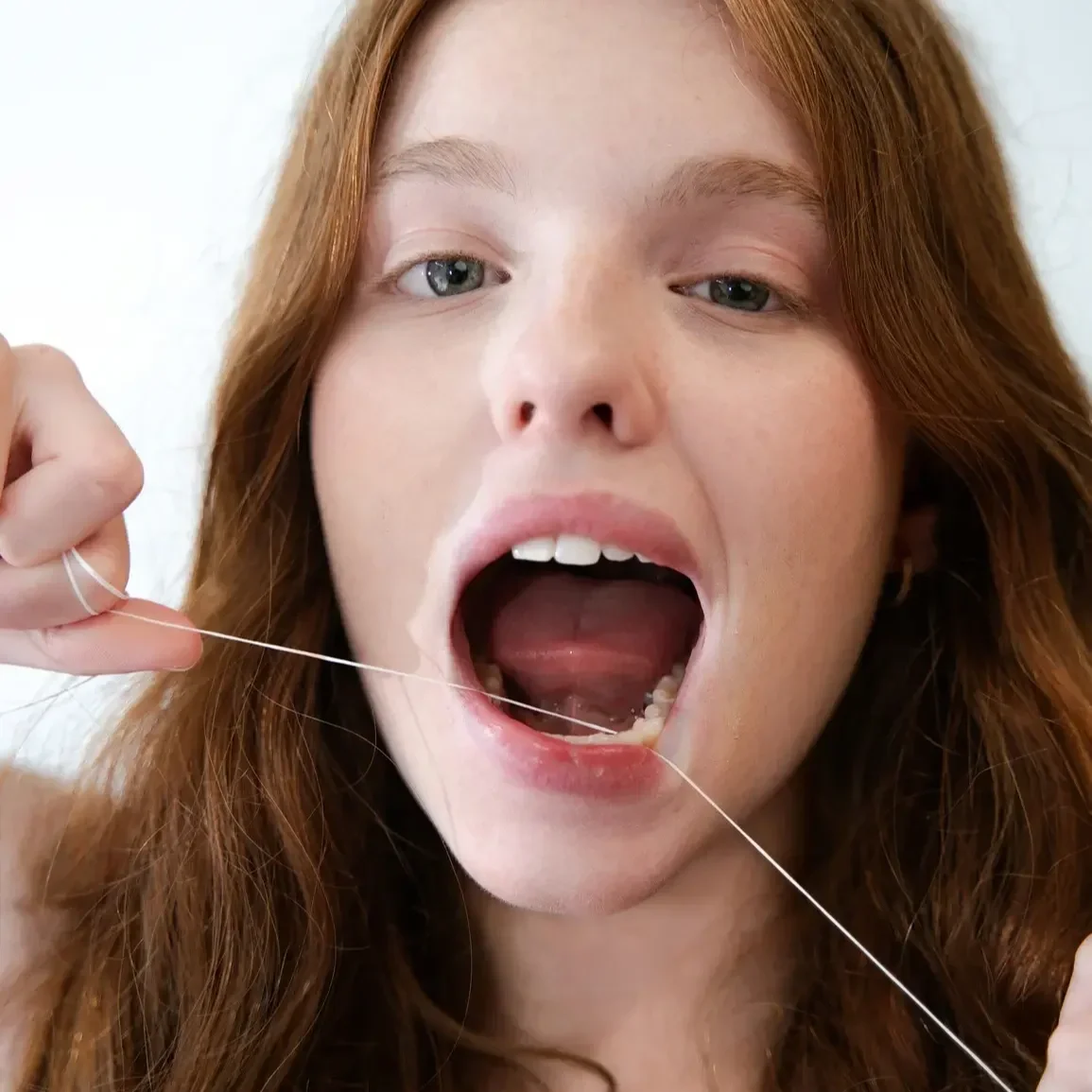 A young woman with red hair and blue eyes opening her mouth wide, holding a piece of white dental floss in one hand, showing her teeth while flossing.