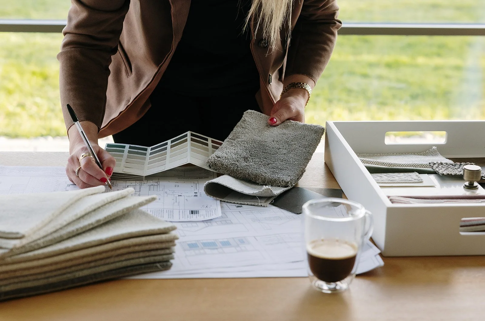 A person working on interior design plans at a wooden desk; they are holding fabric samples and using a color swatch, with a cup of coffee nearby.