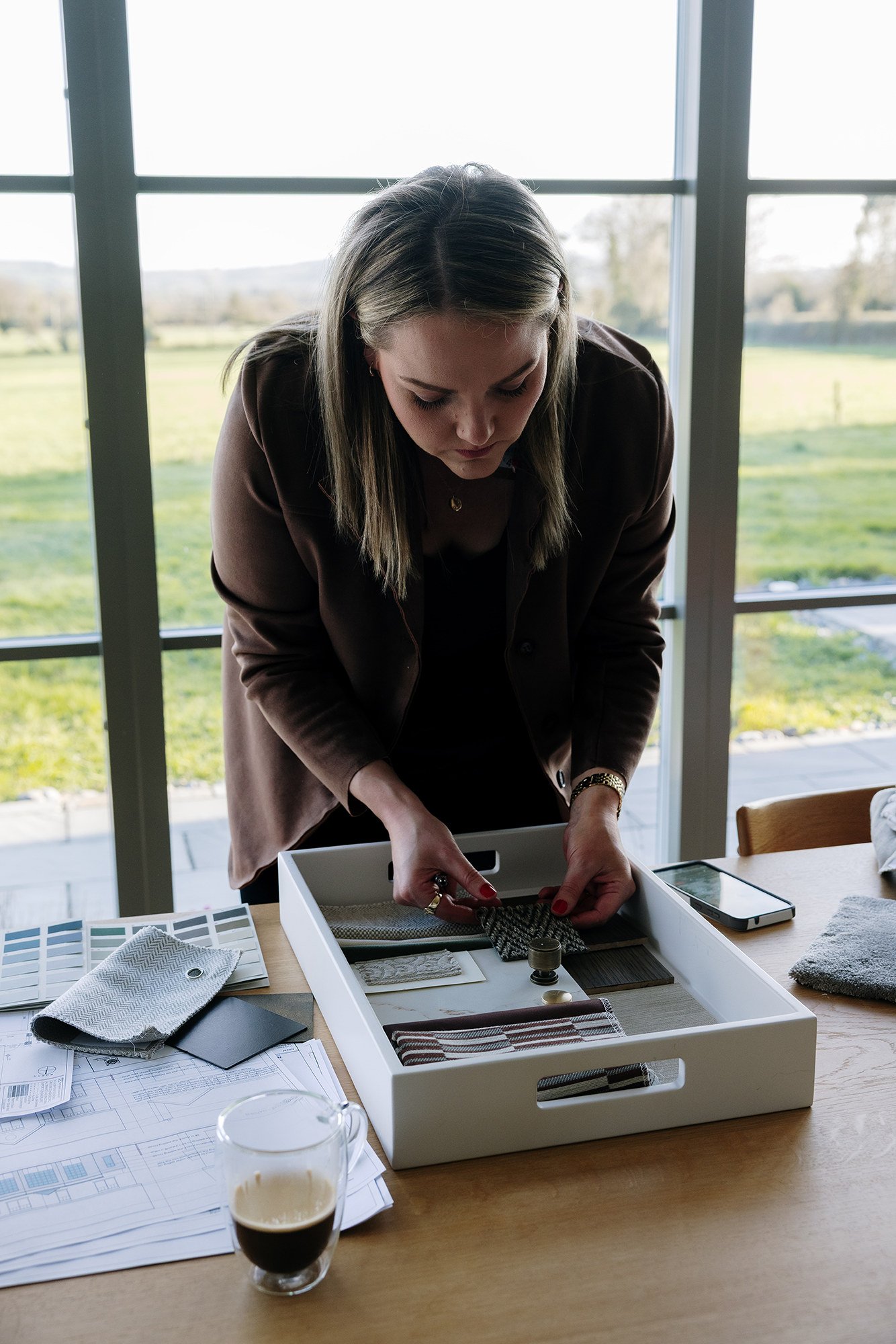 Woman leaning over table with fabric swatches and design plans, near a window with a green landscape outside.