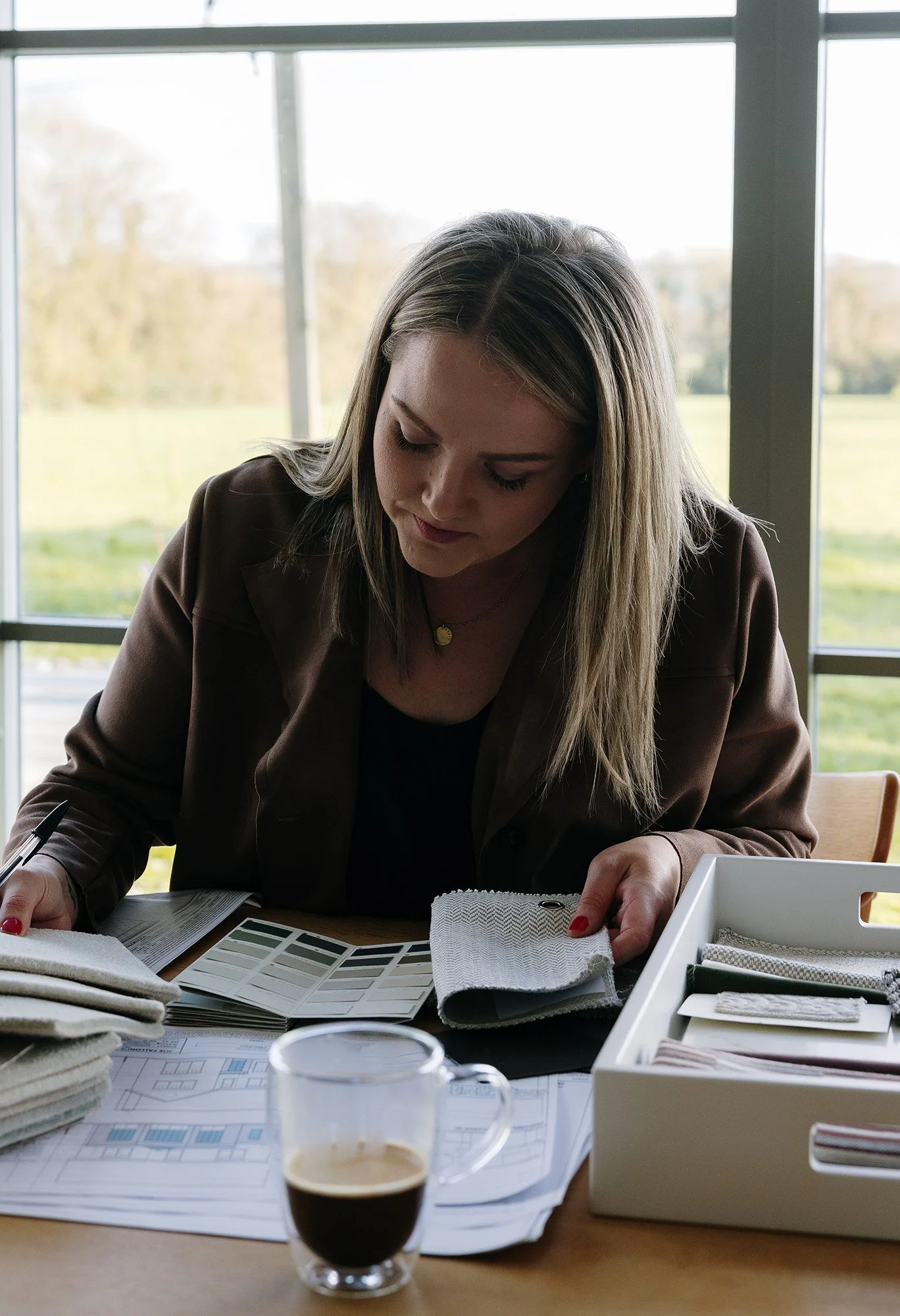Woman looking at fabric samples and color swatches at a work desk with architectural plans and a cup of coffee.
