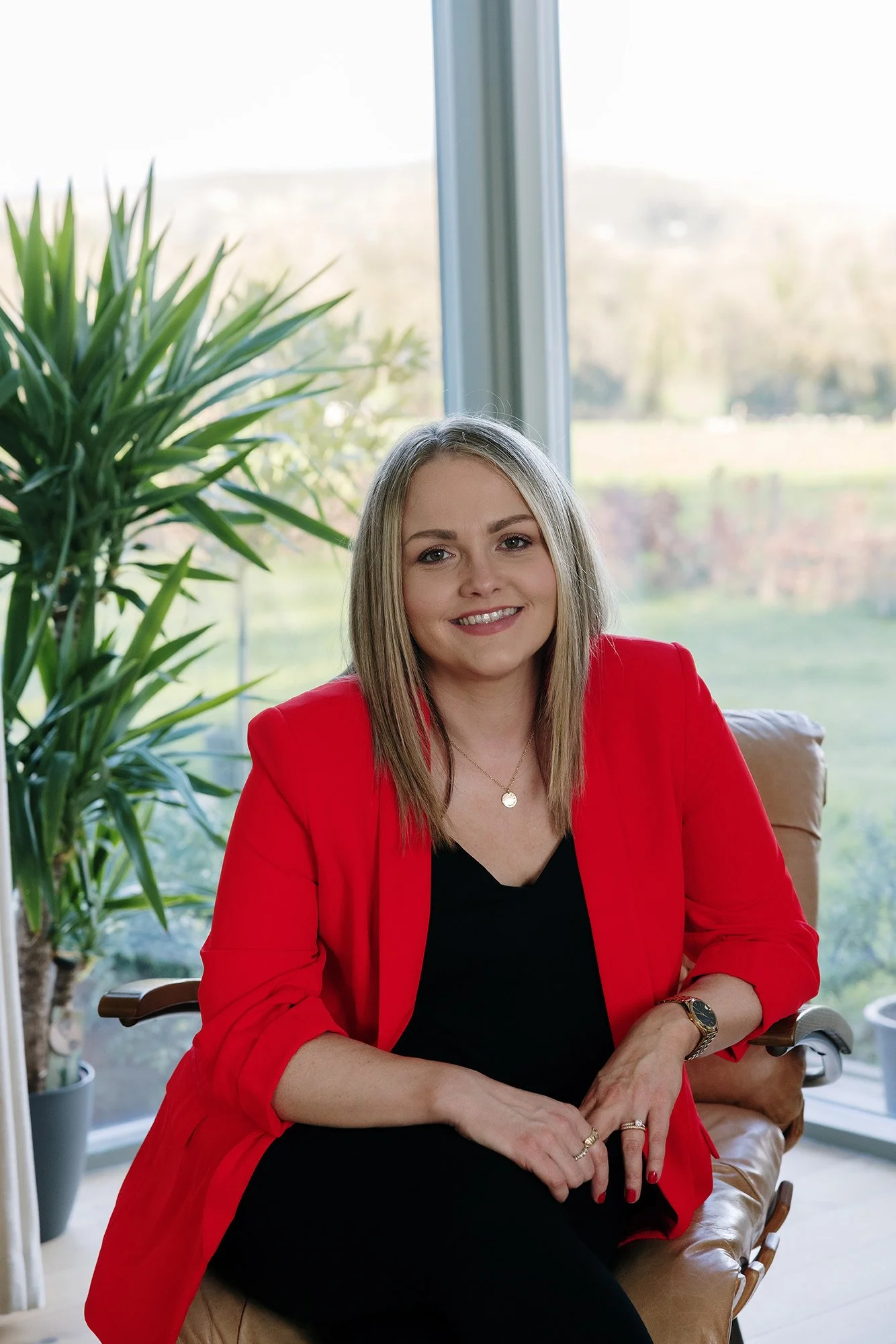 A woman with blonde hair sitting on a brown leather chair, wearing a red blazer and black shirt, smiling at the camera in an office with large windows and a green plant in the background.