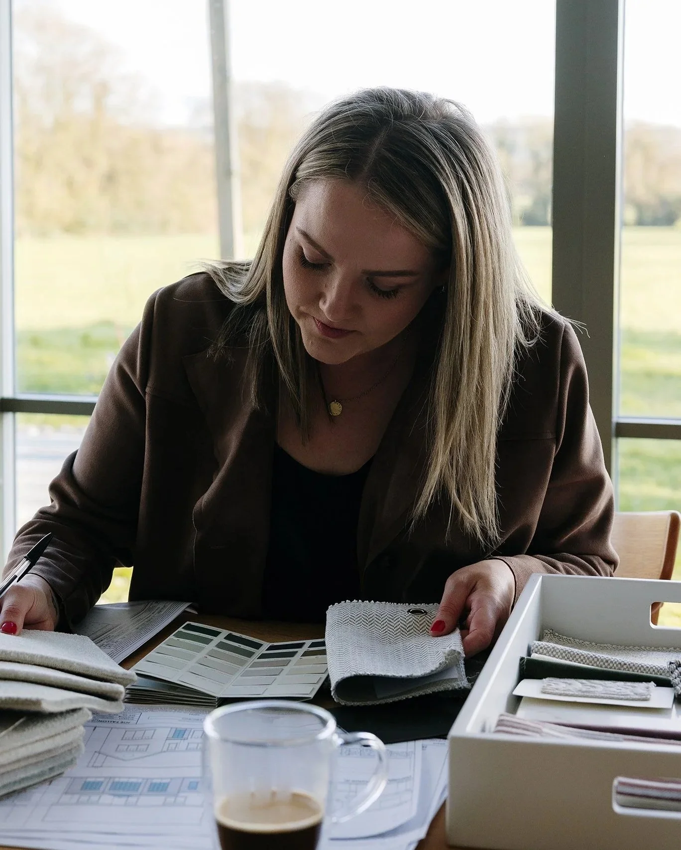 A woman with blonde hair seated at a desk reviewing color swatches and fabric samples, with design documents and a coffee in front of her, during daytime.