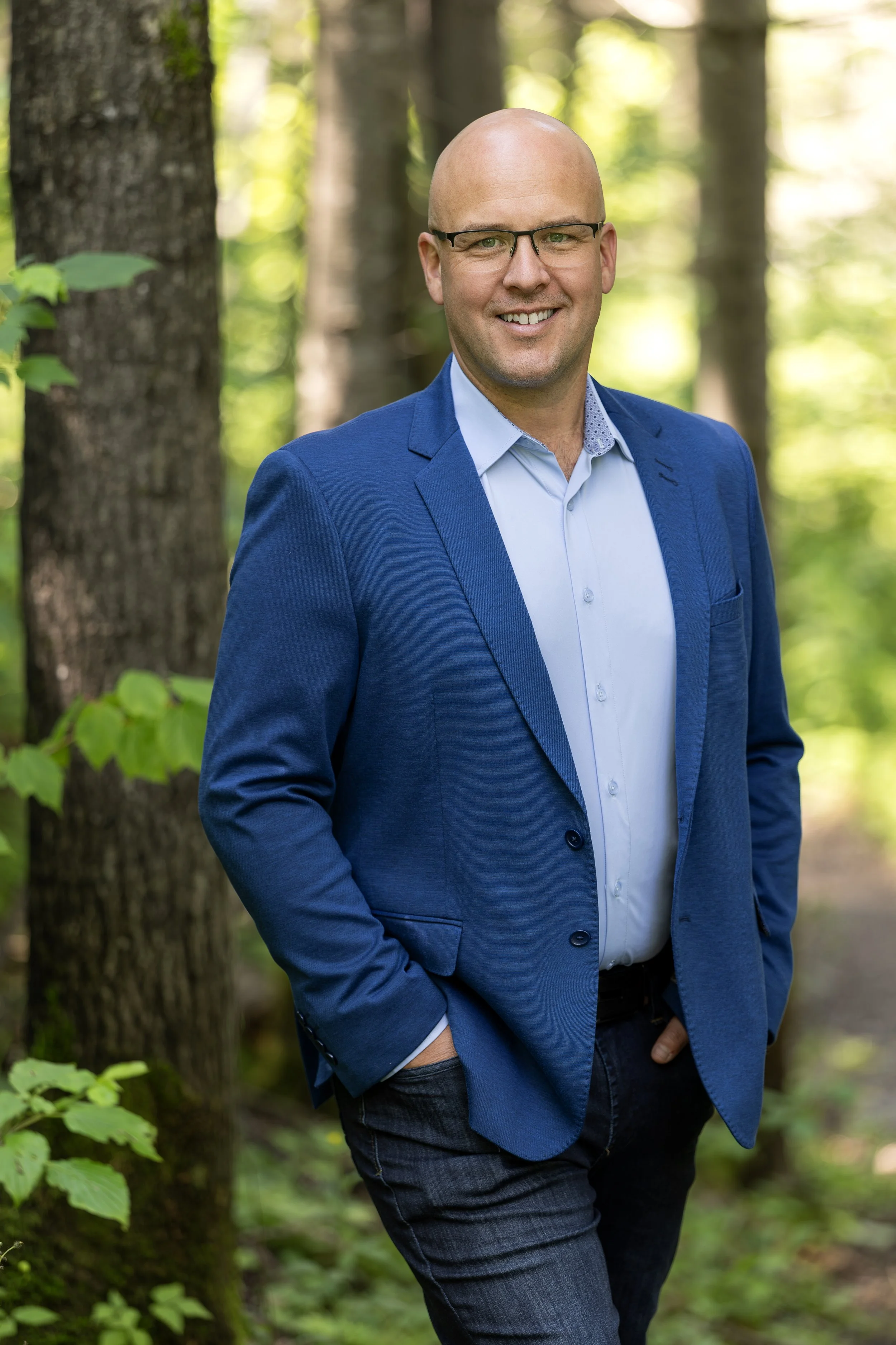 Johnatan Doucet, consultant et fondateur de Mosaïque Stratégie. Un homme souriant portant un costume bleu et des lunettes, debout dans une forêt verte