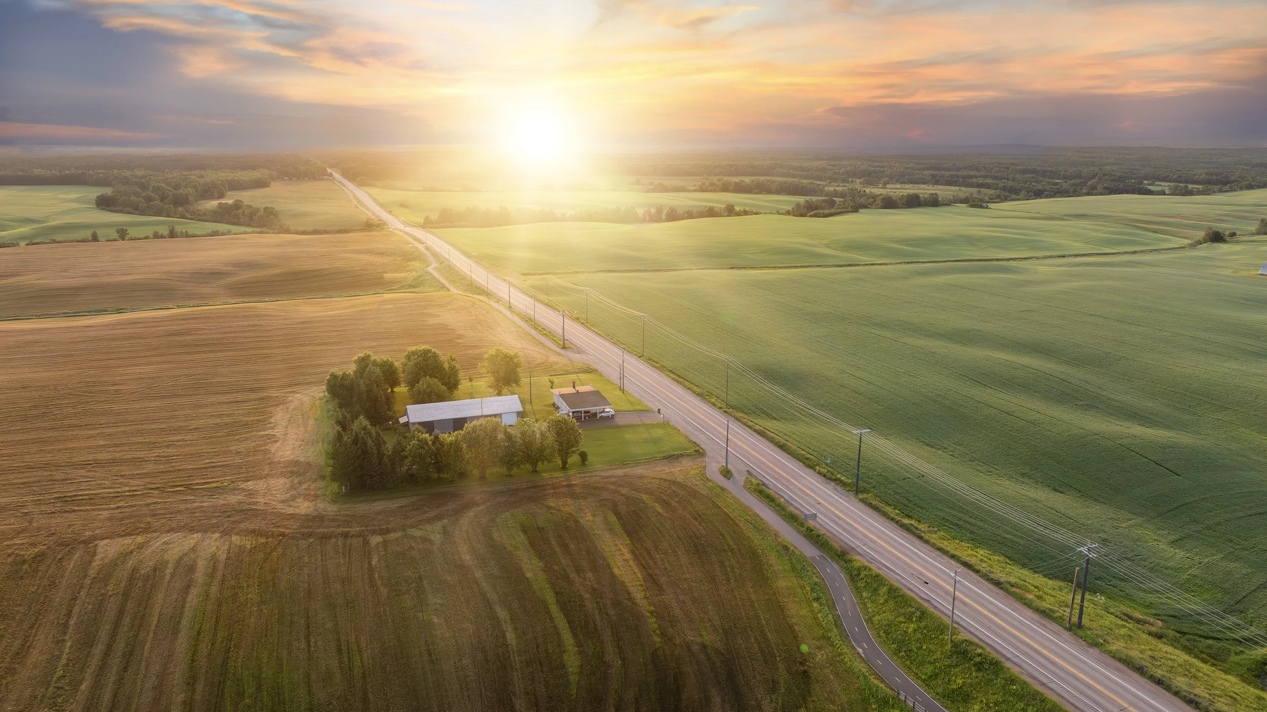 Paysage rural avec champs verts et marrons, une maison entourée d'arbres, une route longeant des lignes électriques, au coucher du soleil.