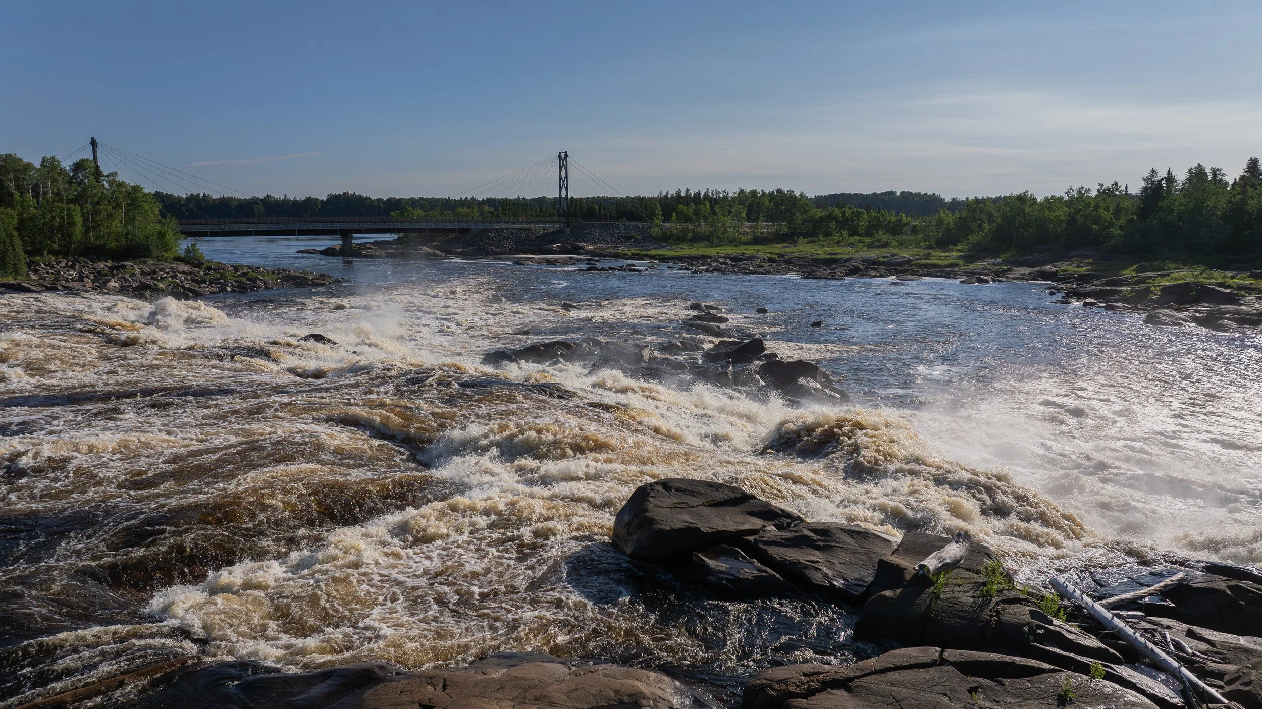 Rivière avec eaux turbulentes et pont suspendu en arrière-plan, entourée de verdure et de ciel bleu.