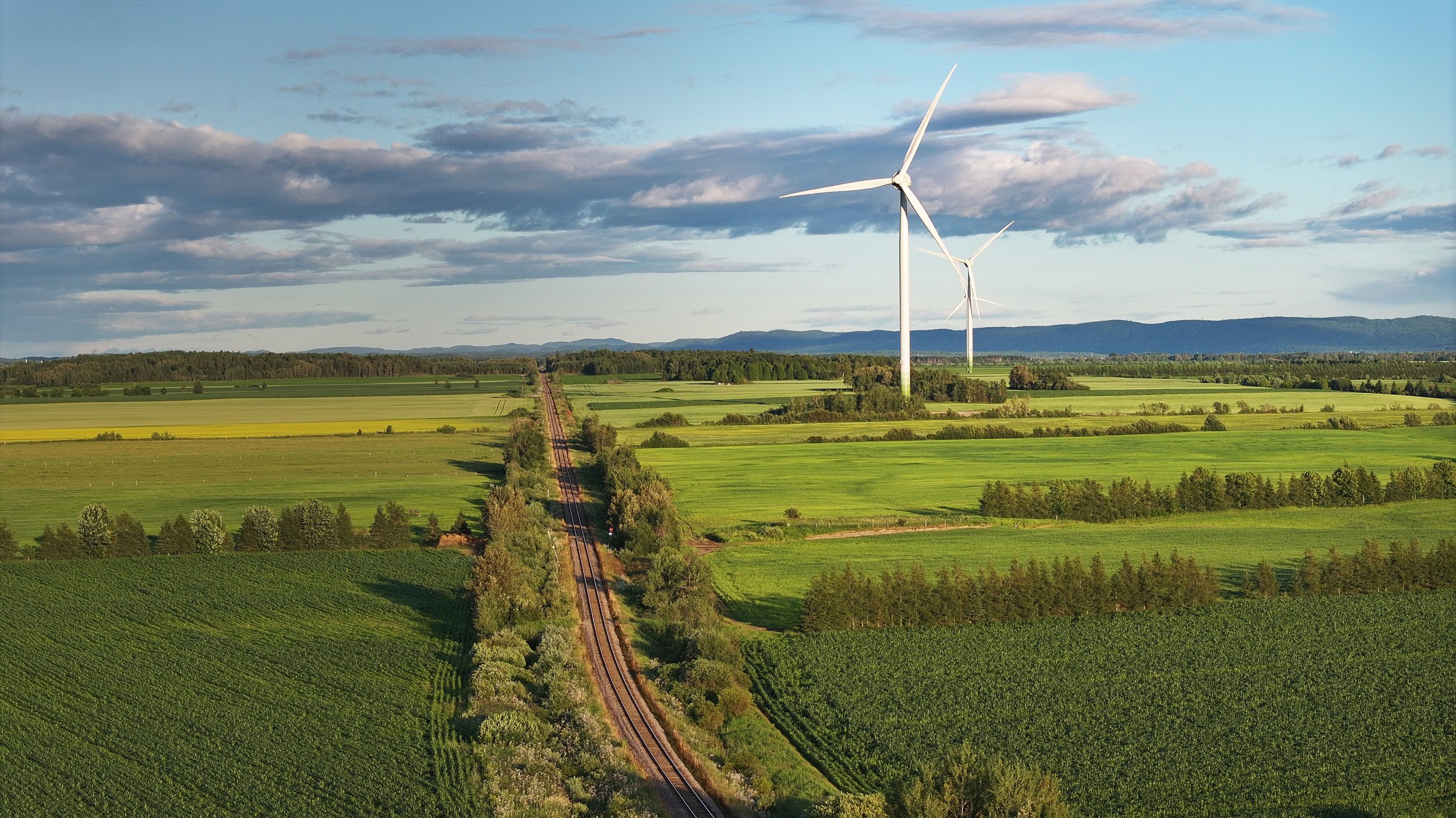 Champs verts avec une voie ferrée, deux éoliennes, ciel partiellement nuageux et montagnes en arrière-plan
