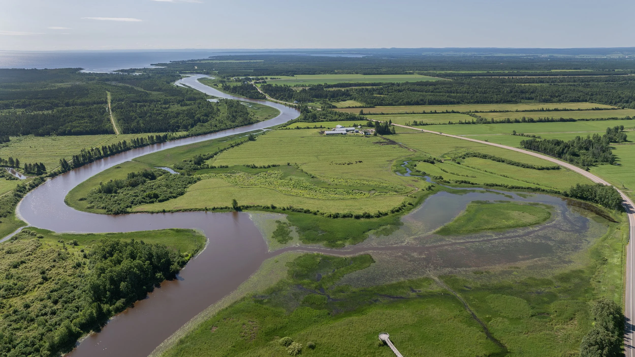 Paysage de champs agricoles, rivières sinueuses, forêts et horizon vers le lac ou la mer, vue aérienne en plein jour.