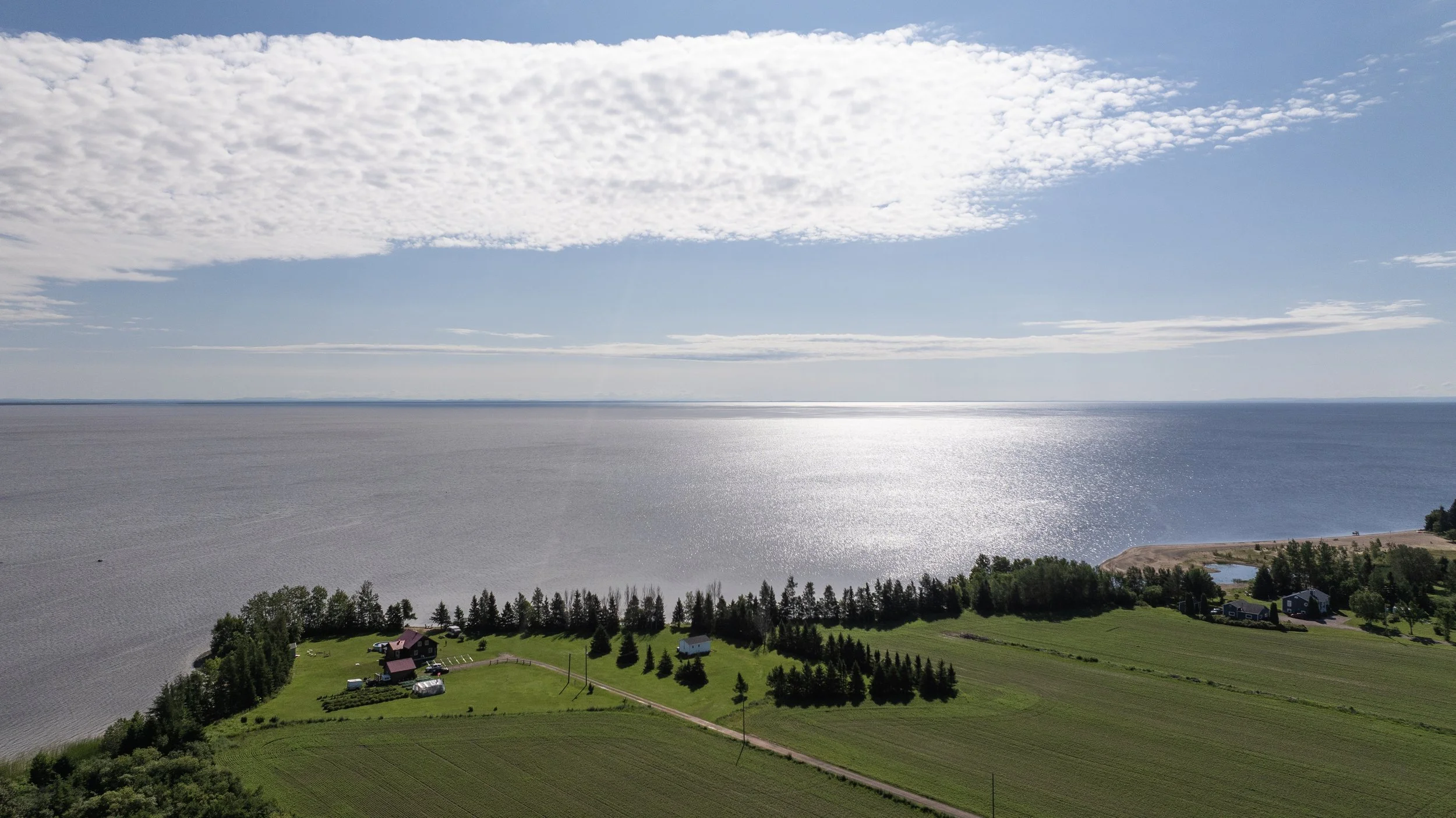 Vue aérienne d'une plage, d'une mer calme, et de champs verts avec quelques maisons, sous un ciel partiellement nuageux.
