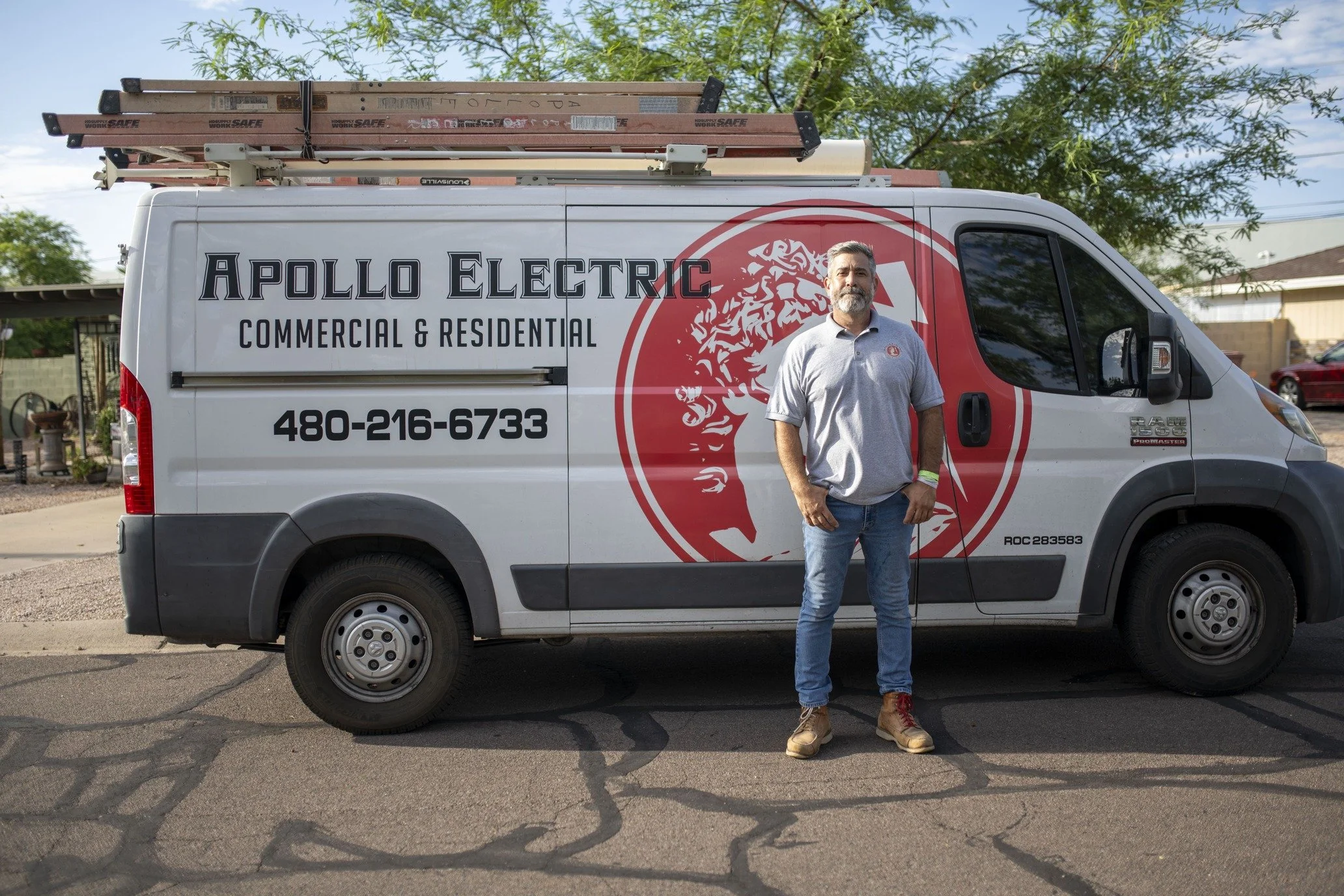 Man standing next to a white Apollo Electric service van with a large red logo and text for commercial and residential electrical work, parked on a cracked street with houses and trees in the background.