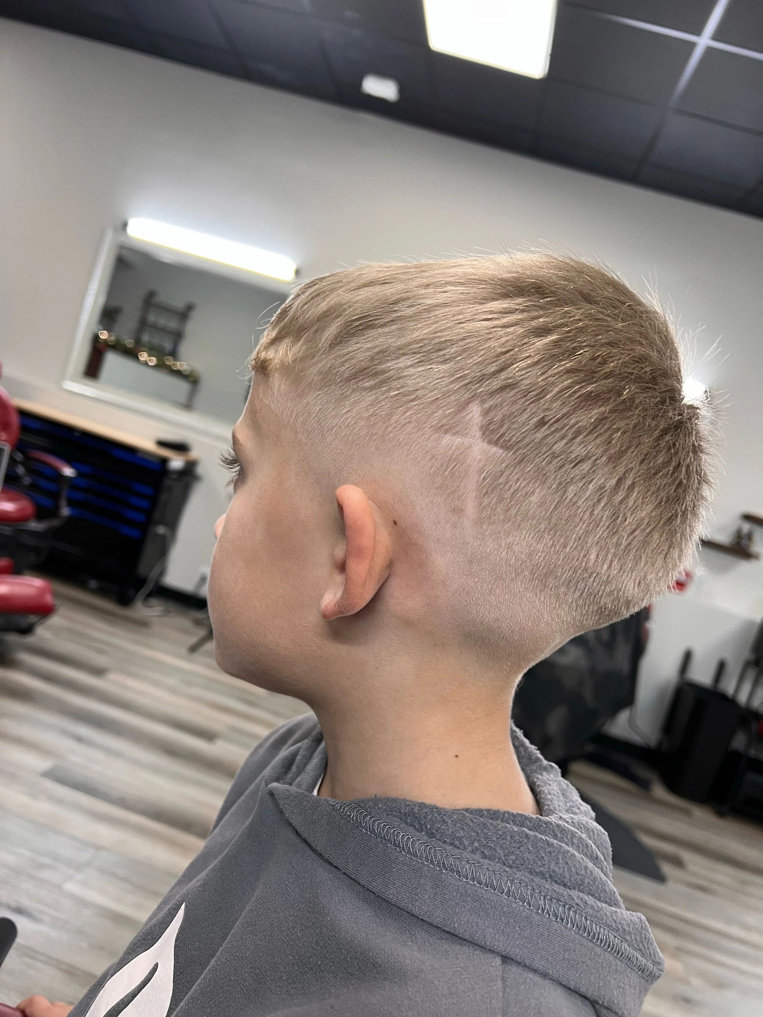 A young boy with a fresh blonde haircut, featuring a fade, in a barbershop with mirrors, chairs, and barber tools in the background.