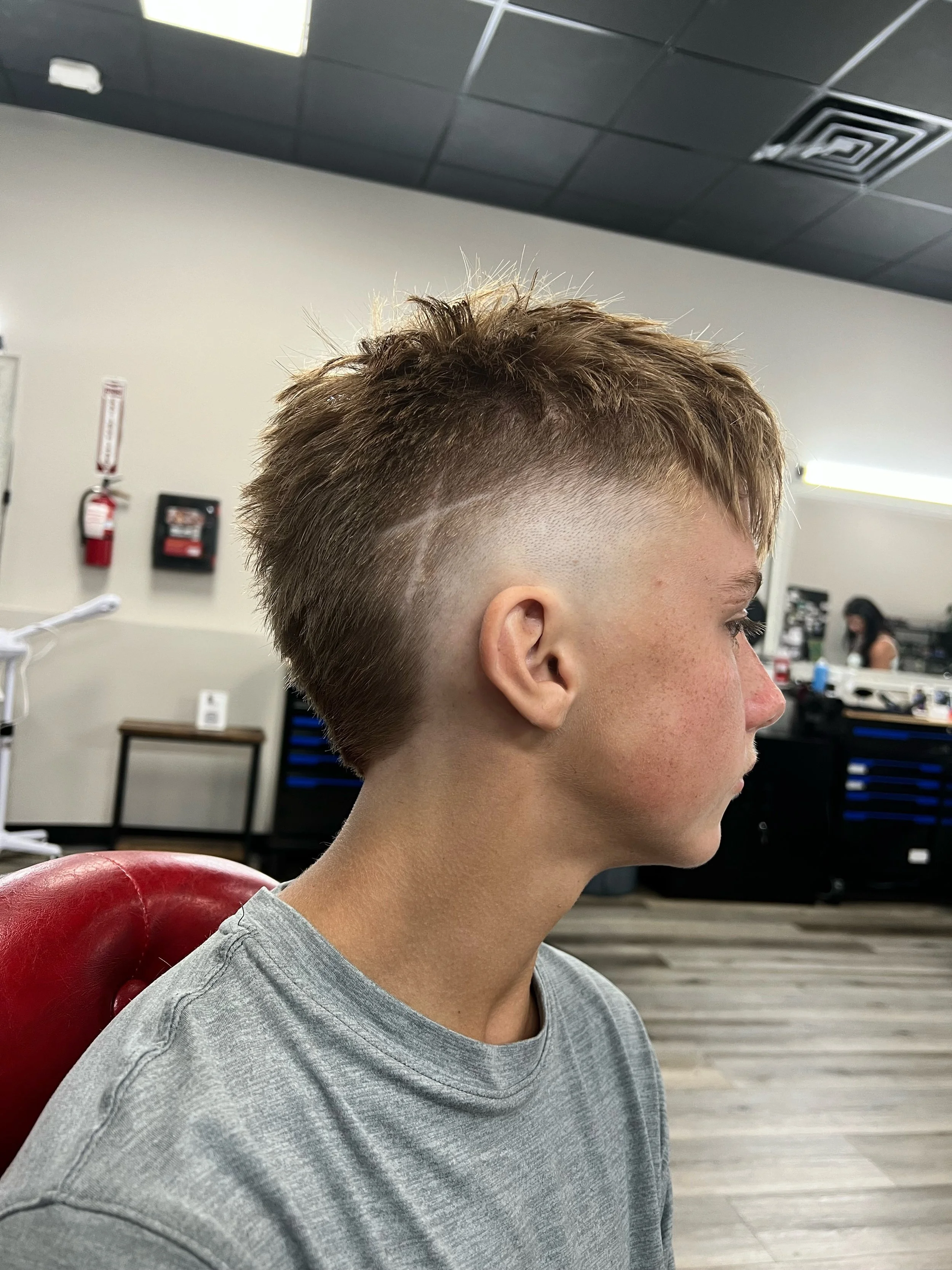 Side view of a person with a modern hairstyle, short on the sides and back with longer textured hair on top, sitting in a salon with a red chair.