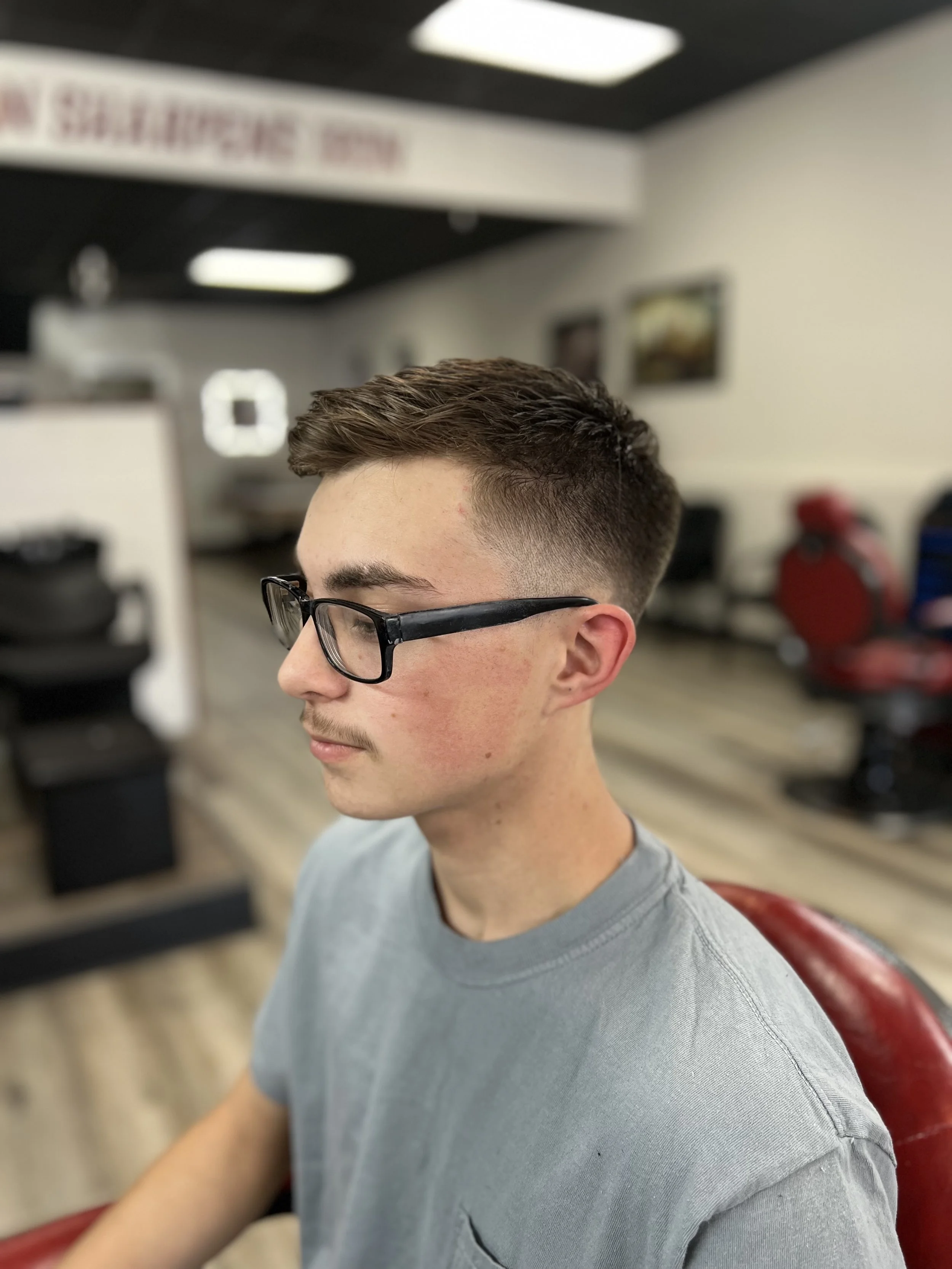 A young man with glasses and a fade haircut sitting in a barbershop.