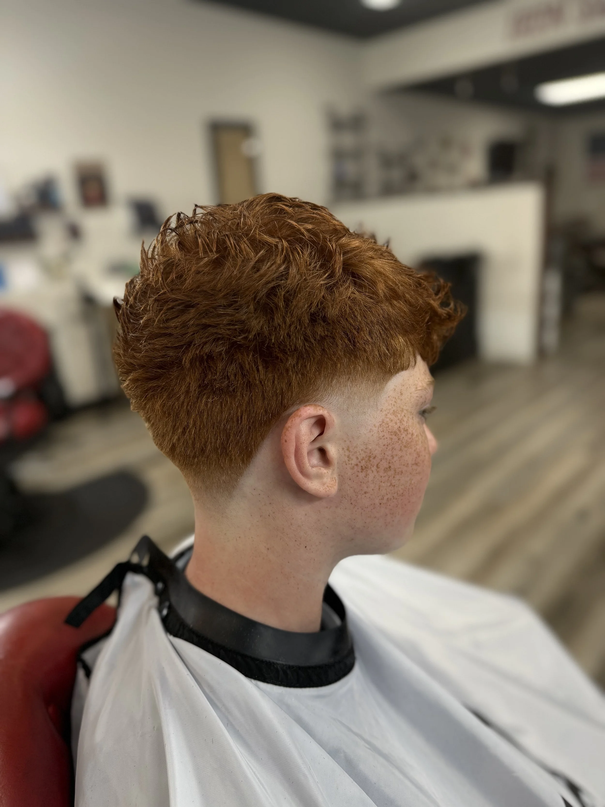 Side view of a young person with short, curly red hair, sitting in a barber's chair, wearing a cape, in a barbershop.