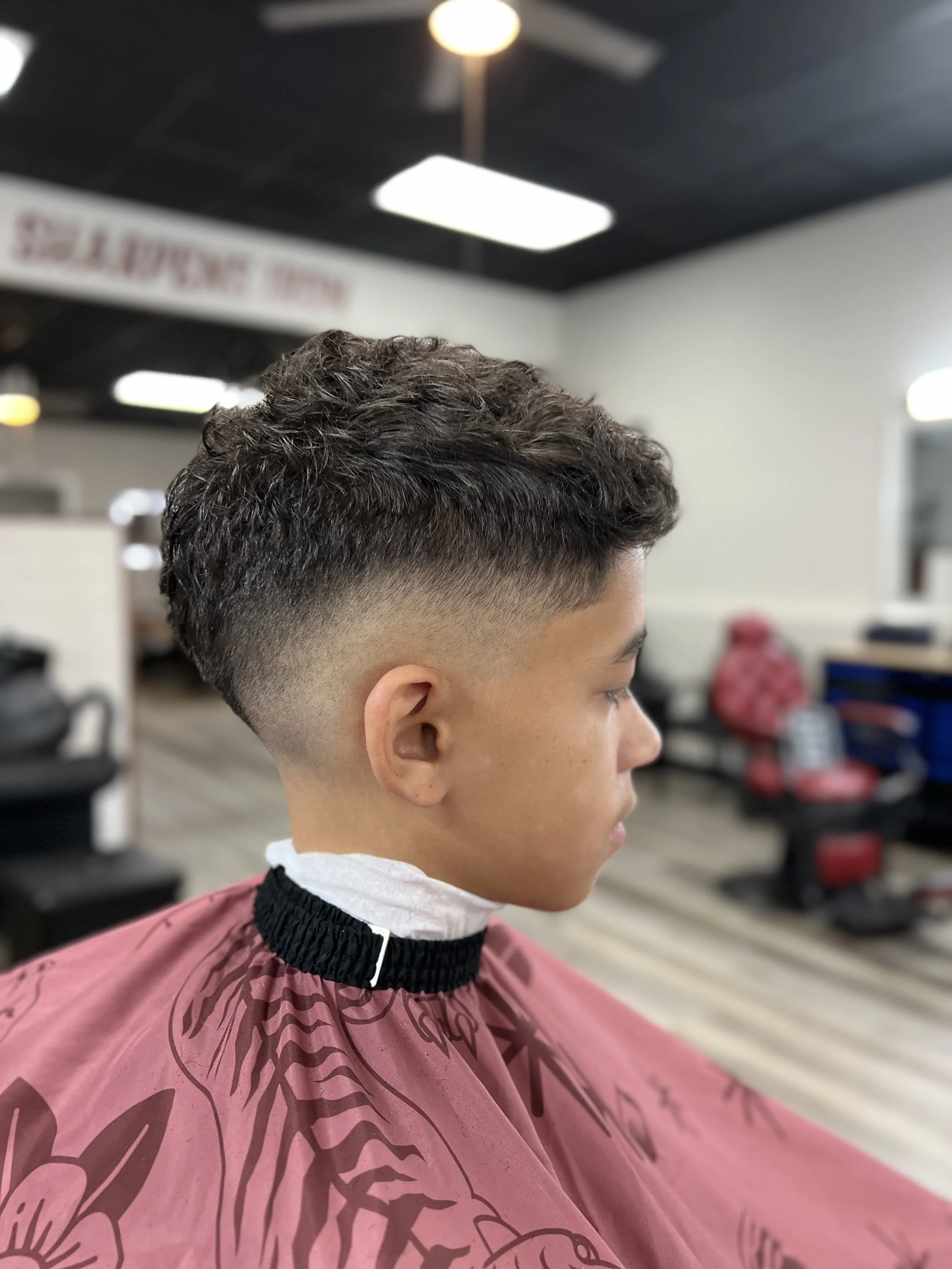 Young man in a barber shop with a fresh curly fade haircut, wearing a pink barber cape with floral patterns.