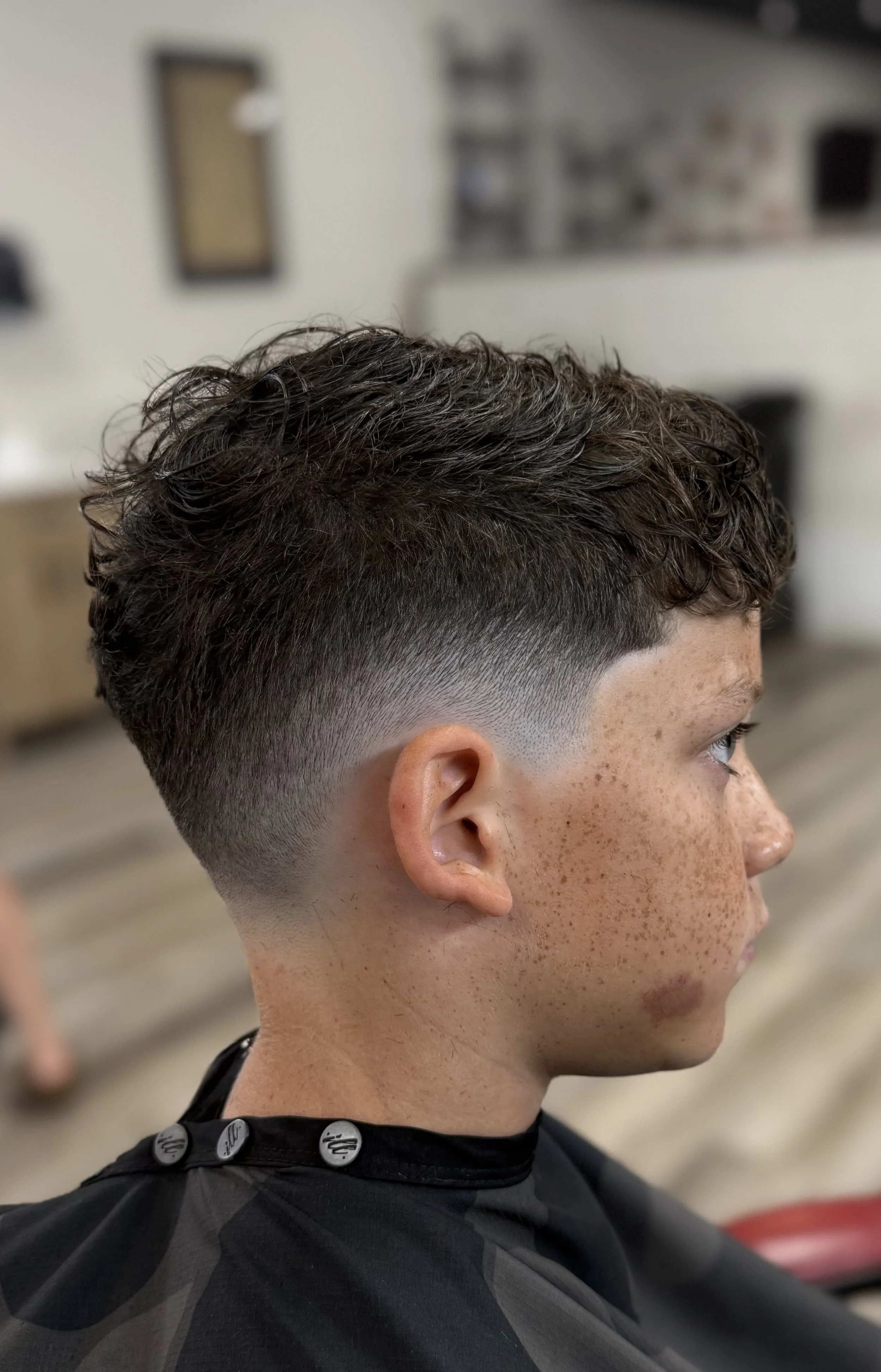 Side view of a young man with a fresh high fade haircut, curly hair on top, and brown freckled skin, sitting in a salon chair.