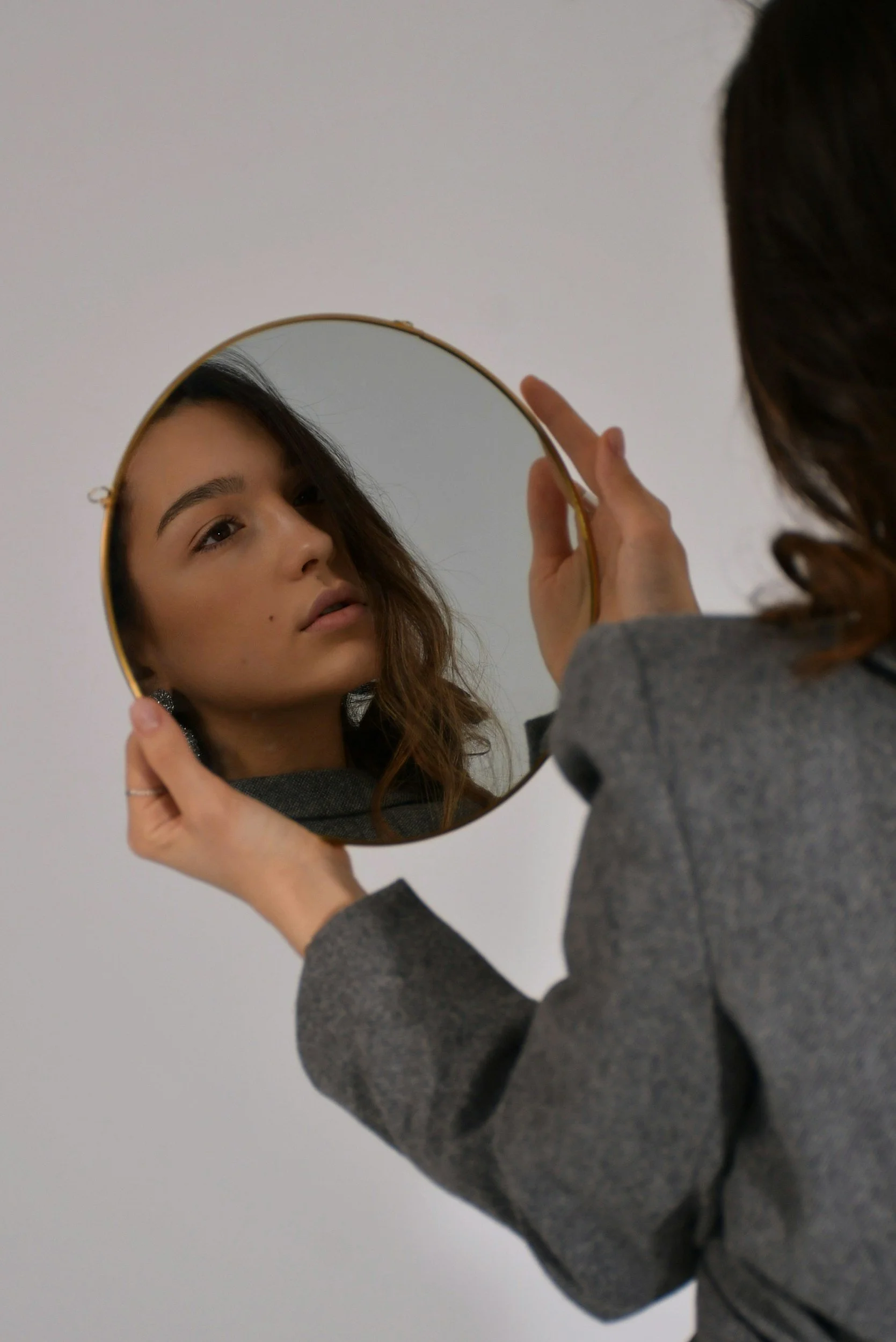 A woman in a gray blazer holds a round mirror, looking at her reflection with a neutral expression against a plain white background.