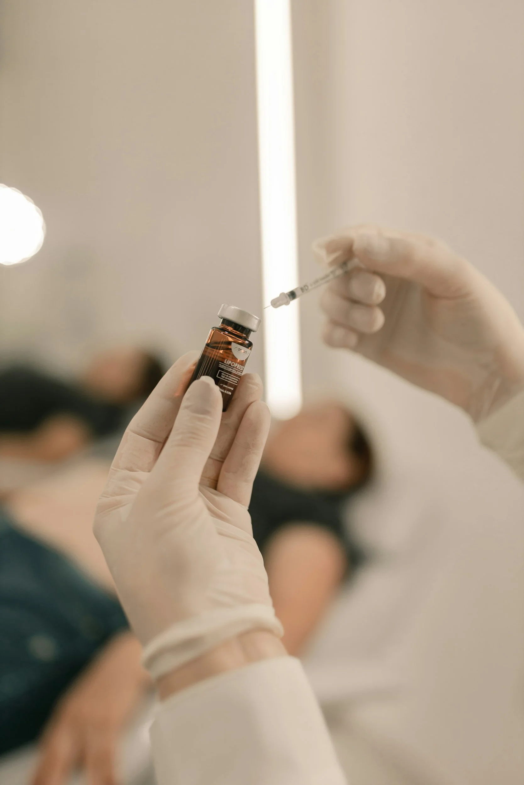 Medical professional wearing gloves preparing a vaccine shot with a syringe and vial, patient lying on bed in background.