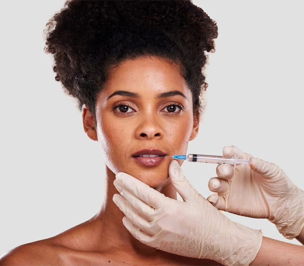 A woman with natural curly hair receives a cosmetic injection in her chin from a healthcare professional wearing gloves.