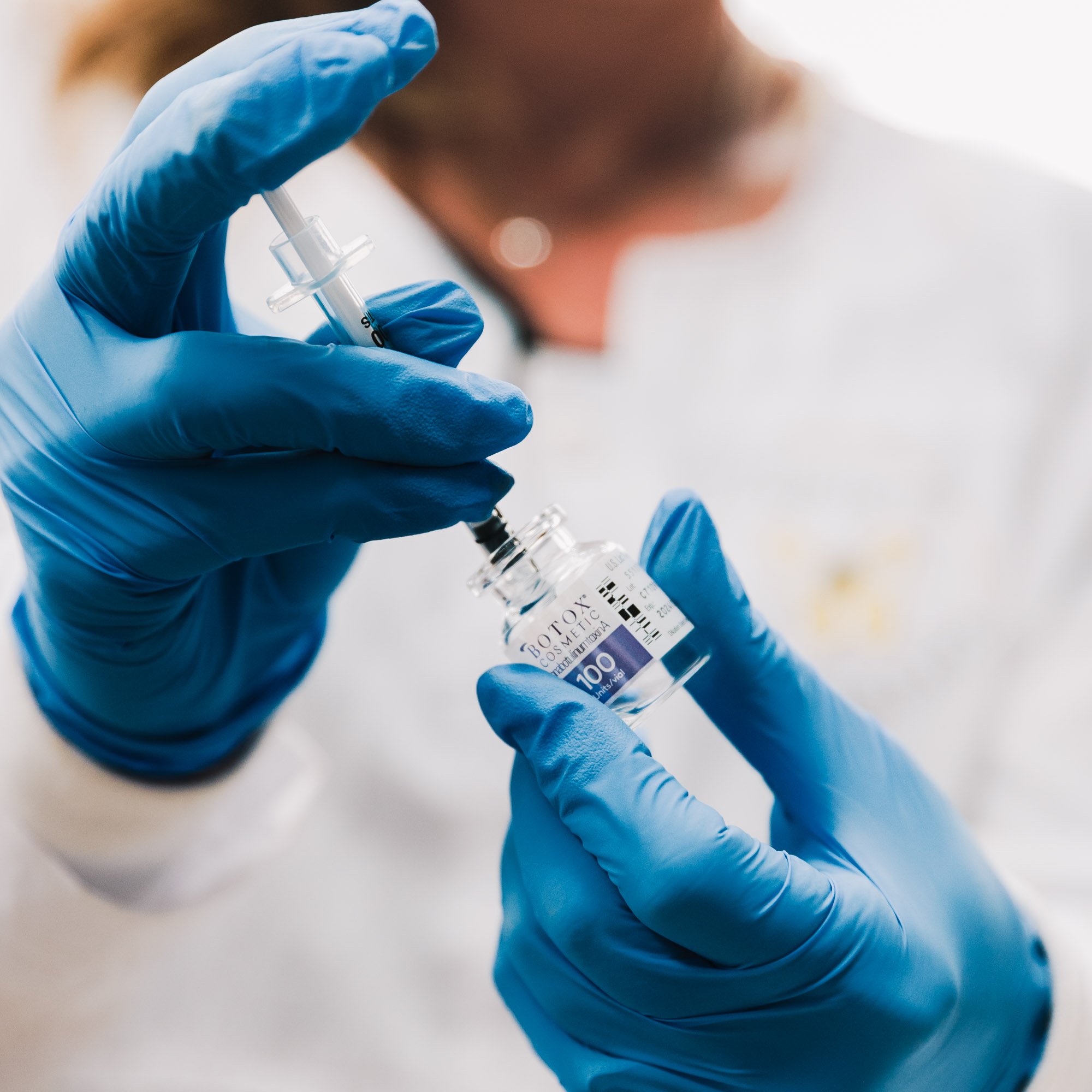 A healthcare professional wearing blue gloves prepares a syringe with a vial of toxin labeled 'Botox Cosmetic' in a clinical setting.