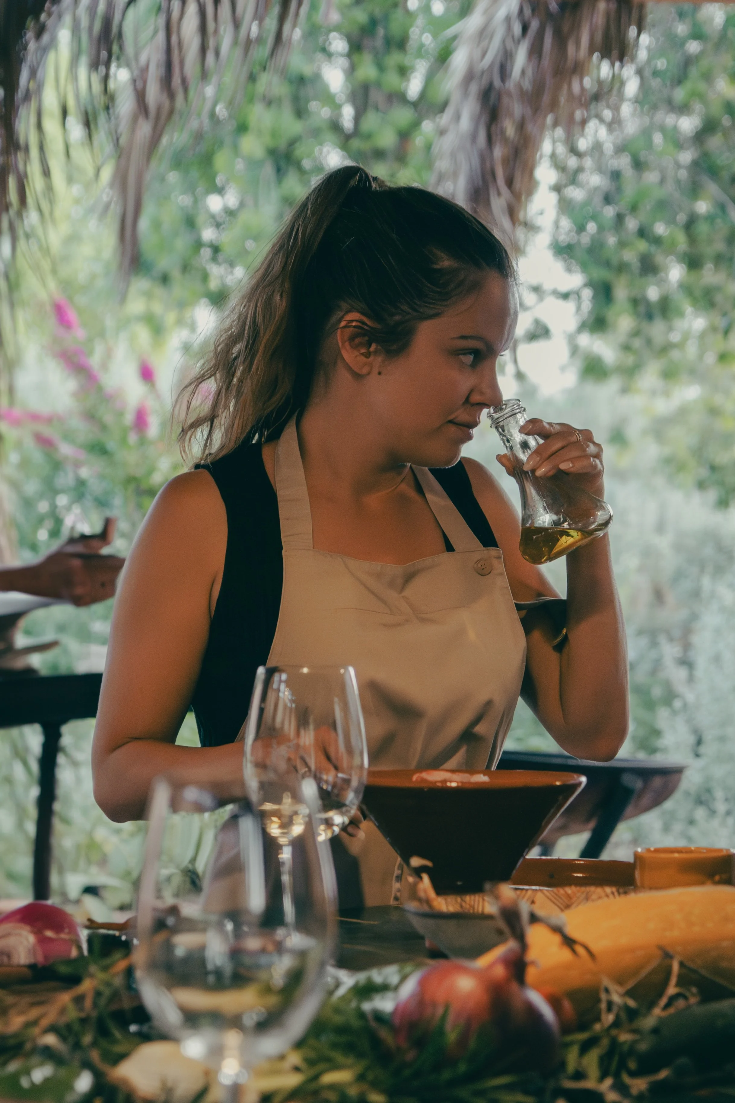 A woman in apron is smelling a bottle of olive oil in a rustic outdoor kitchen setting with trees and greenery in the background. in a hands-on Moroccan cooking class