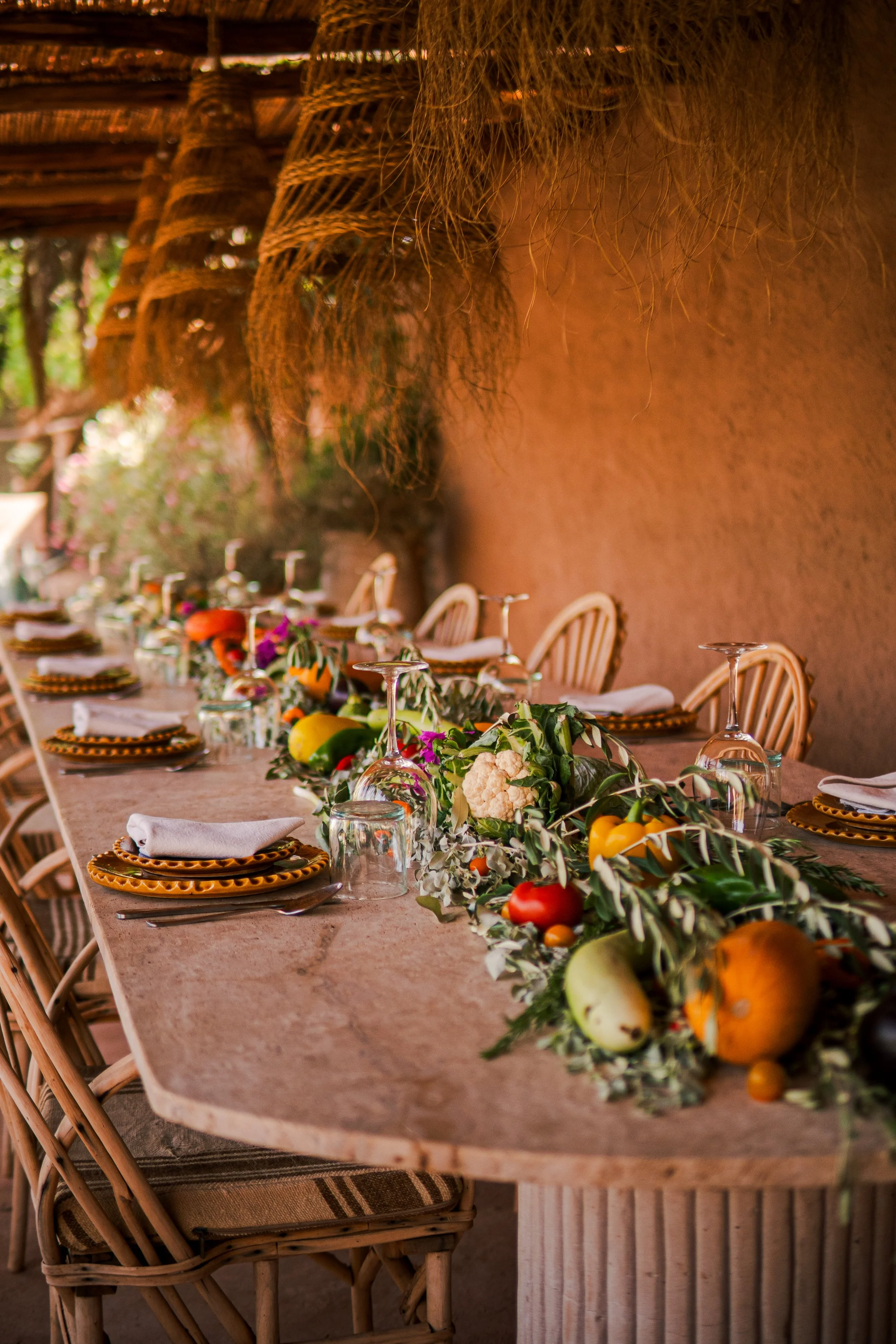 A long dining table decorated with a colorful floral and vegetables centerpieces set under a thatched roof with a rustic, outdoor ambiance in a cooking class in Marrakech,