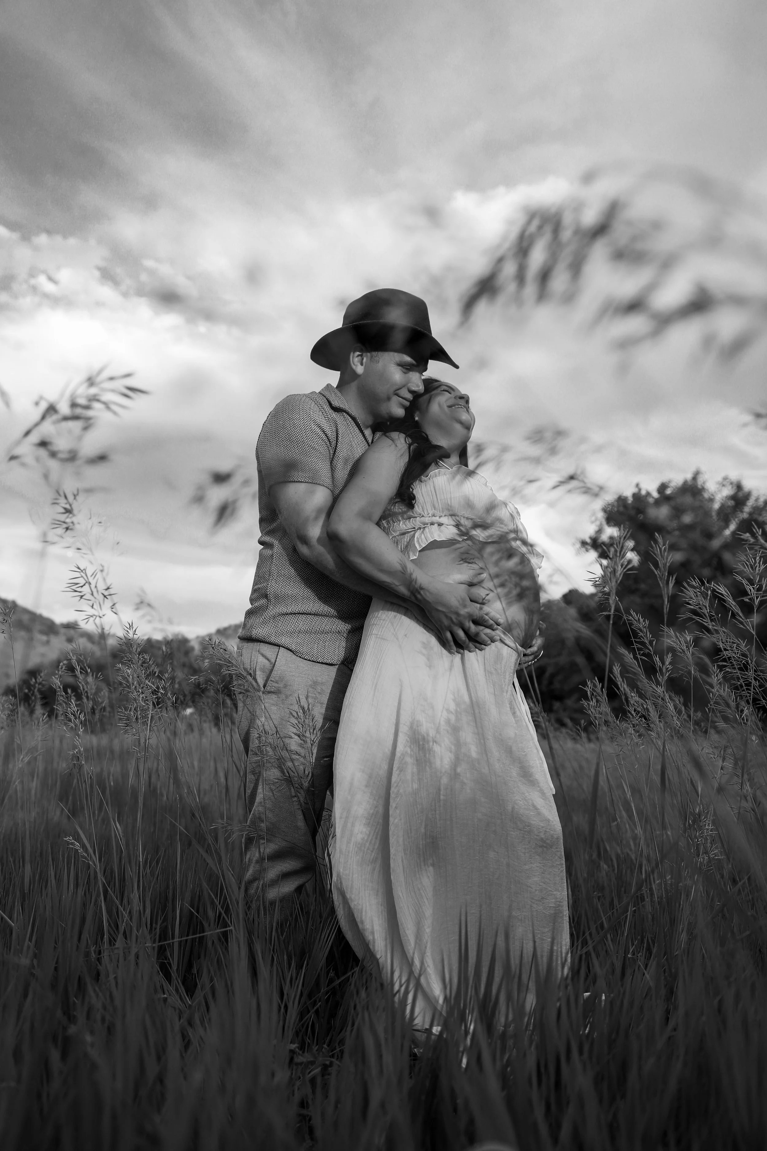 A couple stands in a grassy field, embracing and smiling. The man wears a hat and casual clothing, while the woman is pregnant, wearing a light dress. The sky is partly cloudy, creating a scenic outdoor setting.