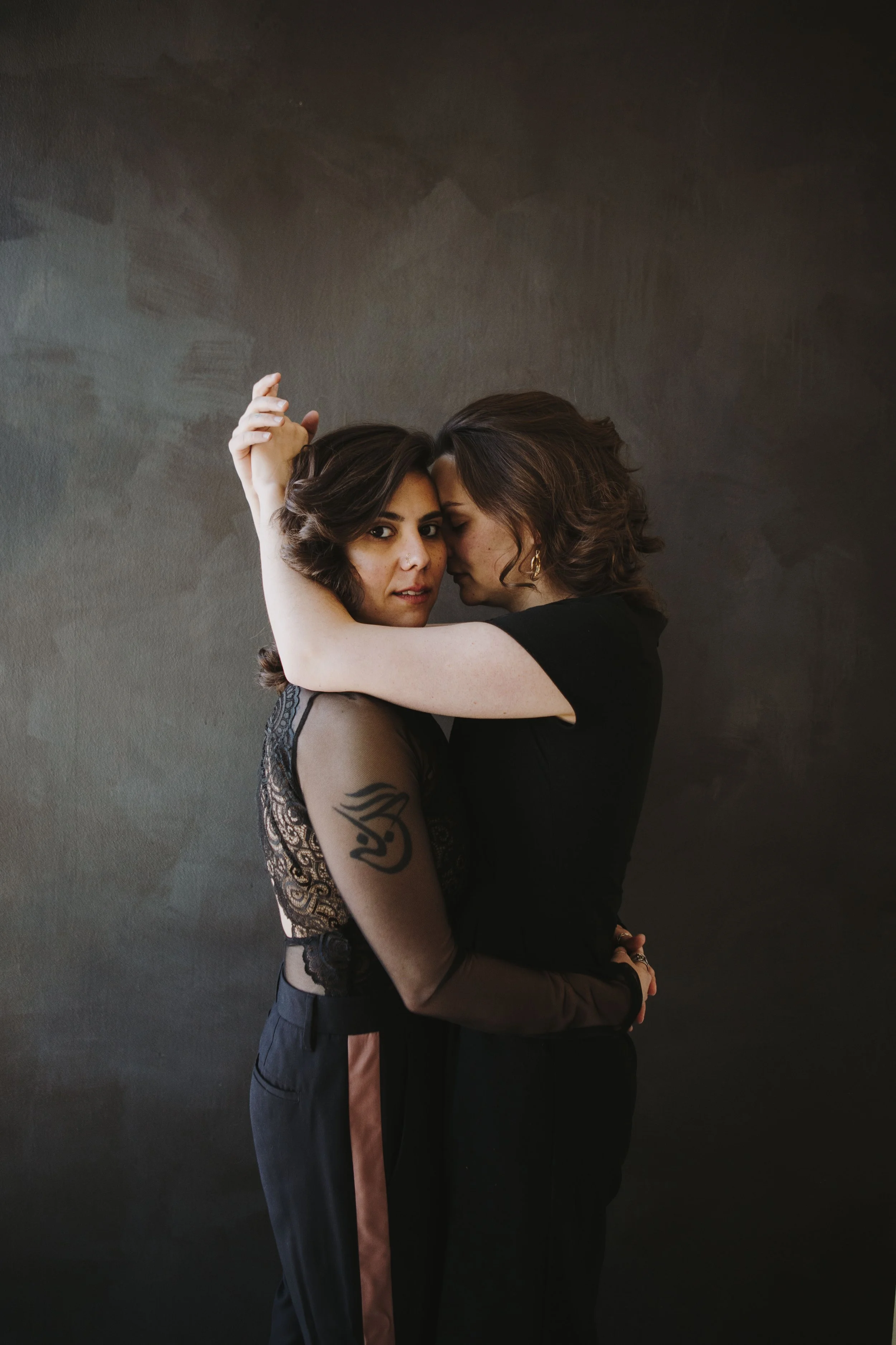 Two women embrace each other closely against a dark background, one with dark hair and the other with light brown hair, both dressed in black.