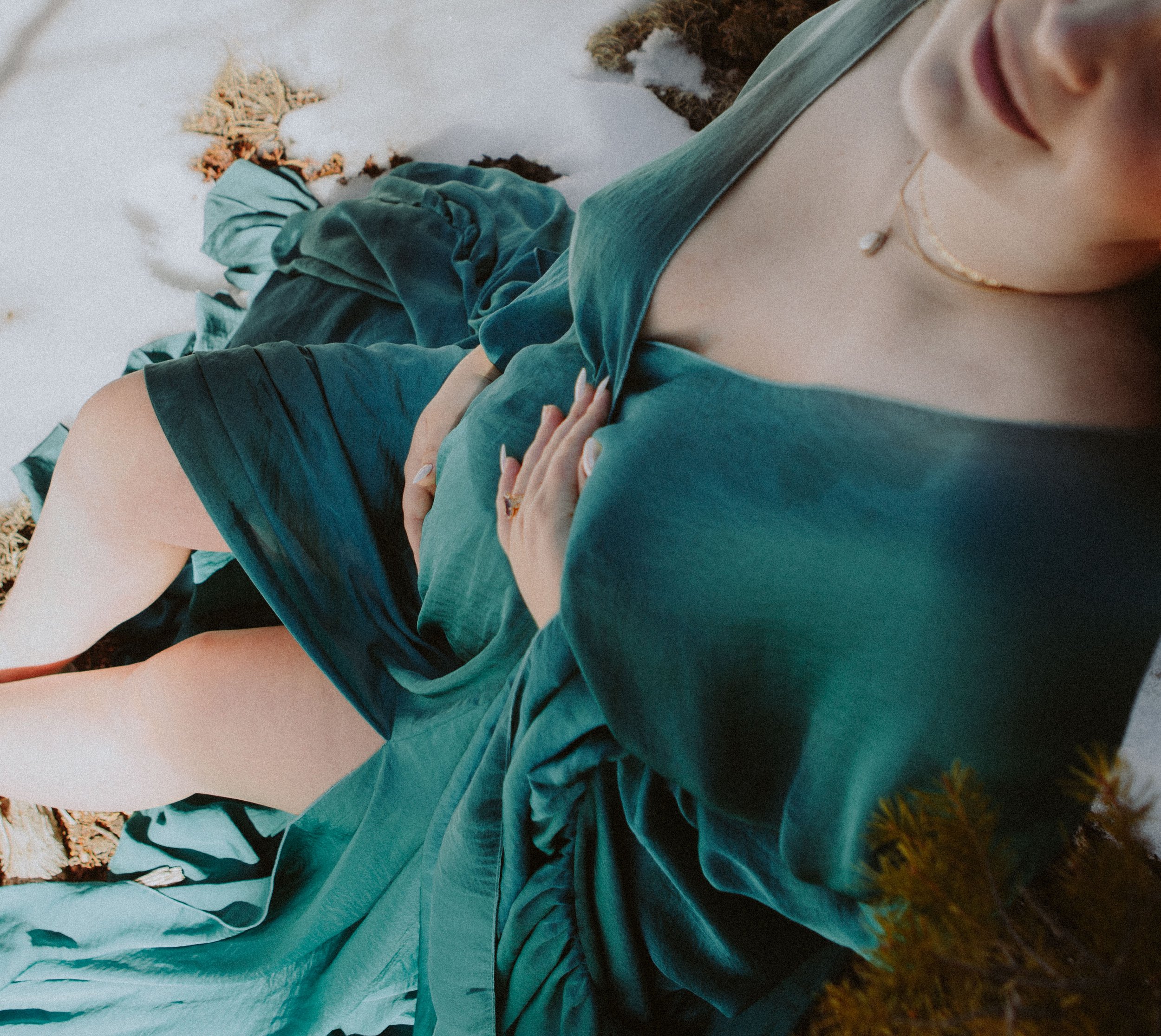 Person in a teal dress sitting on the ground outdoors, with snow and patches of earth visible around.