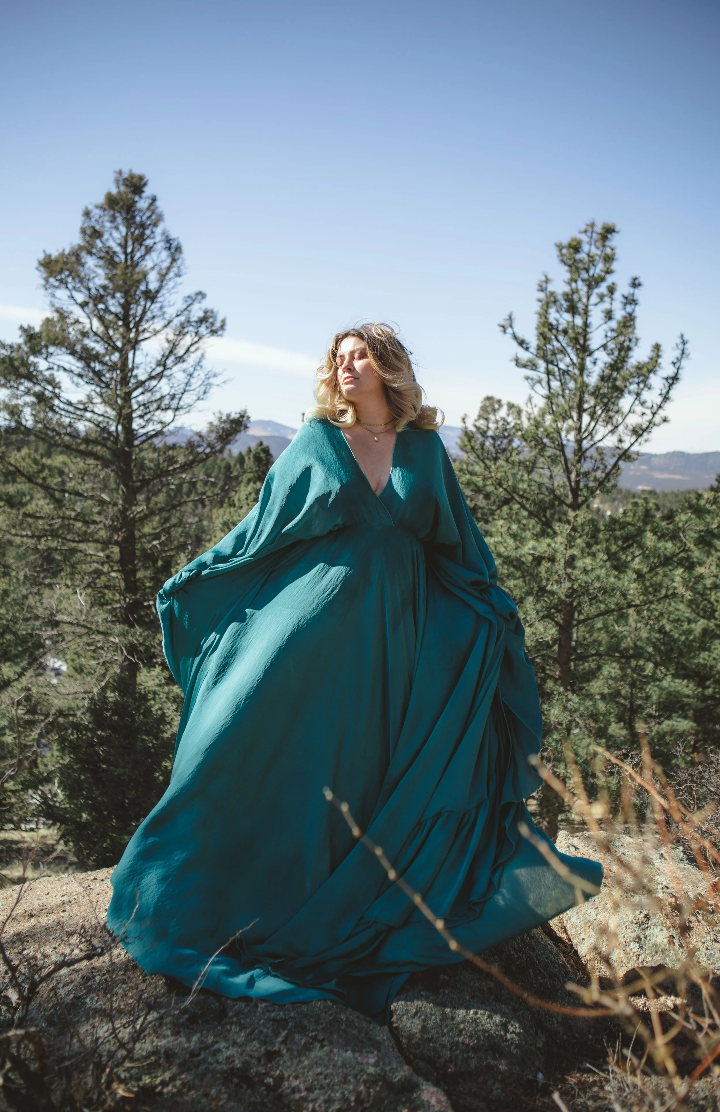 A woman in a flowing, teal dress standing outdoors on rocks with pine trees and mountains in the background under a blue sky.
