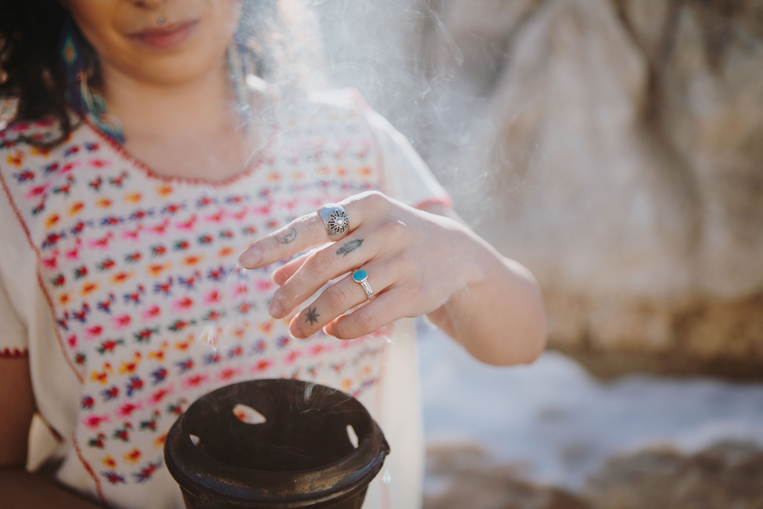 A person with various rings on their fingers, holding burning herbs over a small fire outdoors. The person is wearing a white shirt with a colorful pattern.