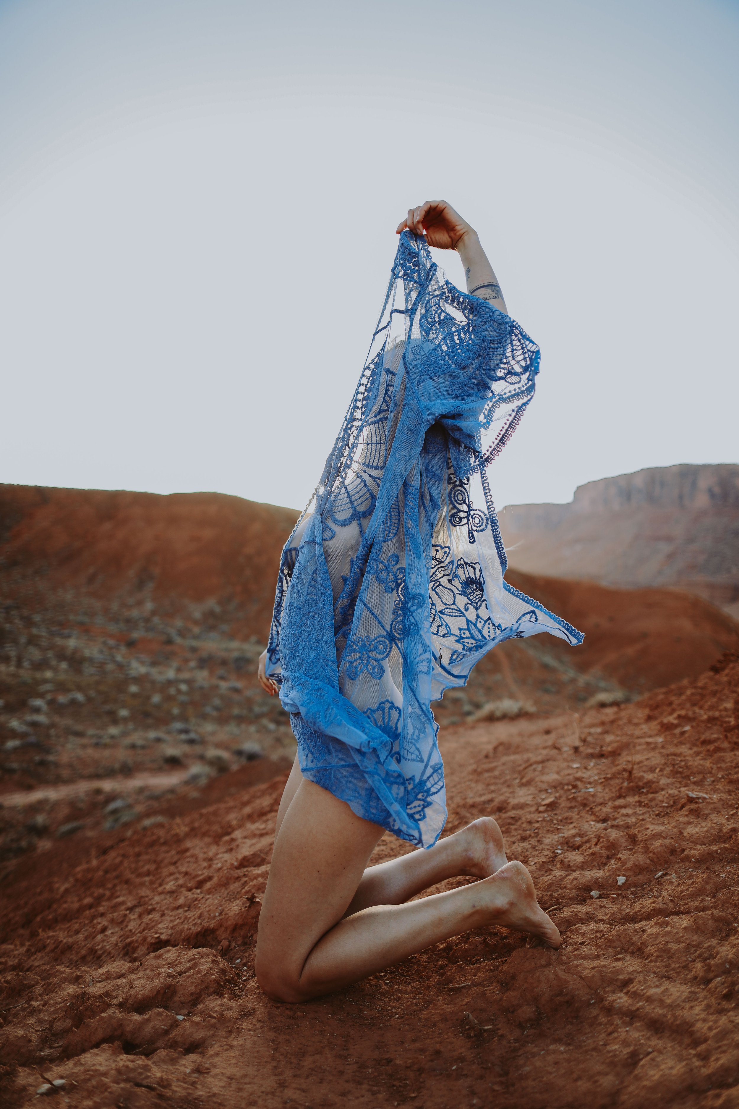 Person kneeling on red desert ground with blue embroidered sheer fabric draped over their head and body, holding the fabric up with one hand, mountain landscape in the background during sunset.