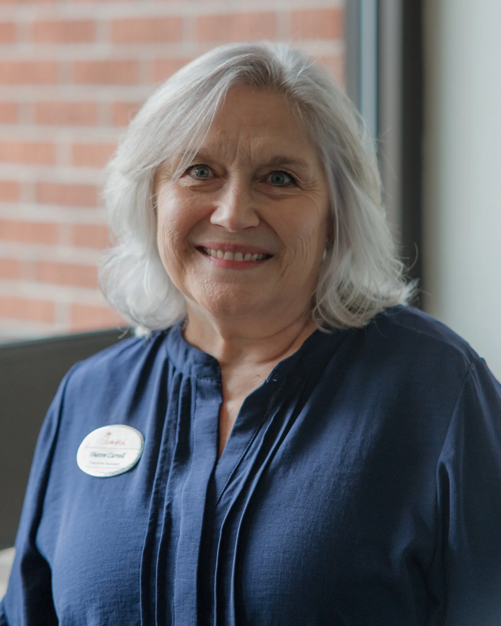 Close-up of a smiling senior woman with gray hair wearing a dark blue uniform and a name tag, standing indoors in front of a brick wall.