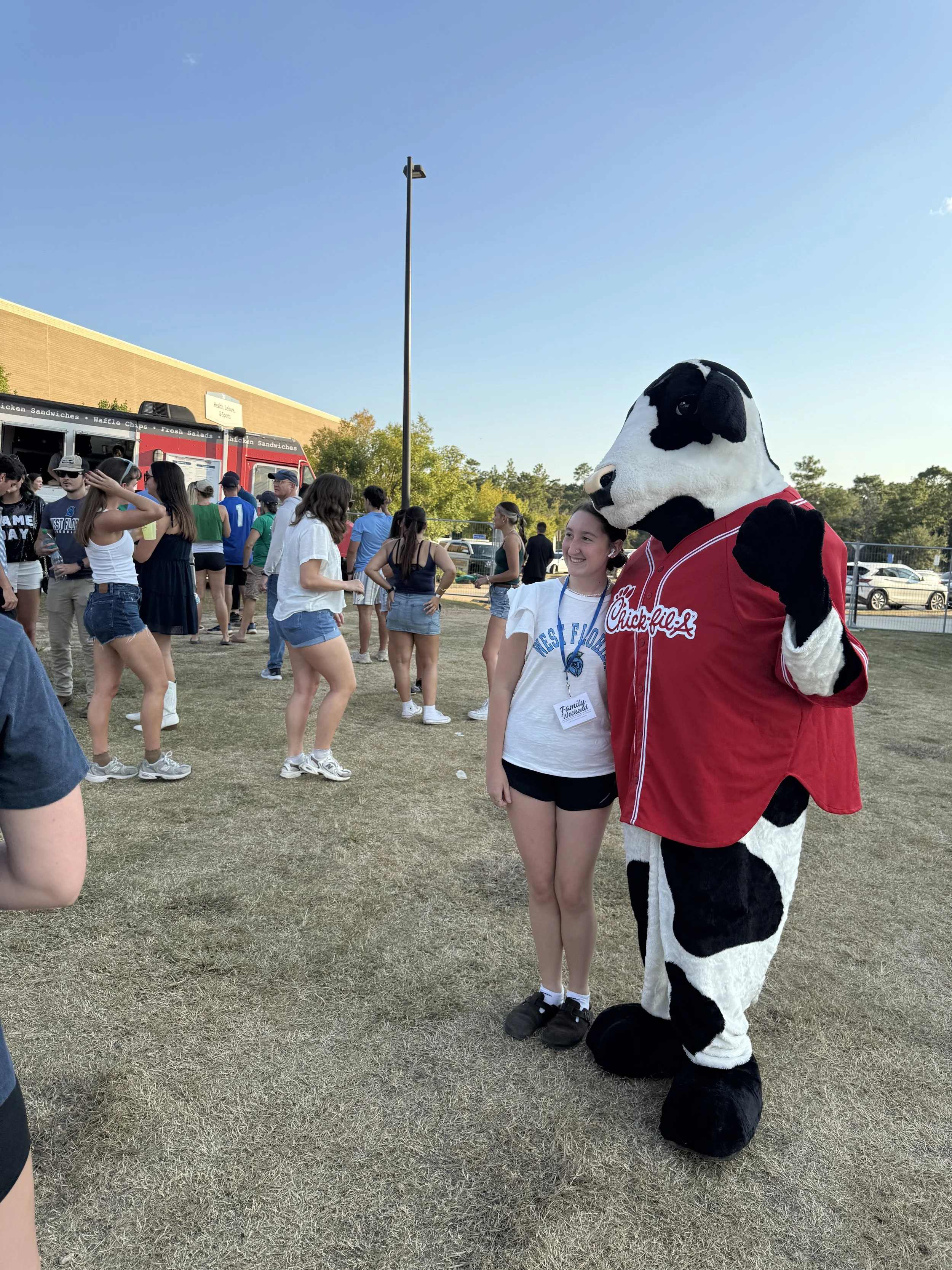 A person in a Chick-fil-A cow costume posing with a girl at an outdoor event with a food truck and a crowd of people in the background.