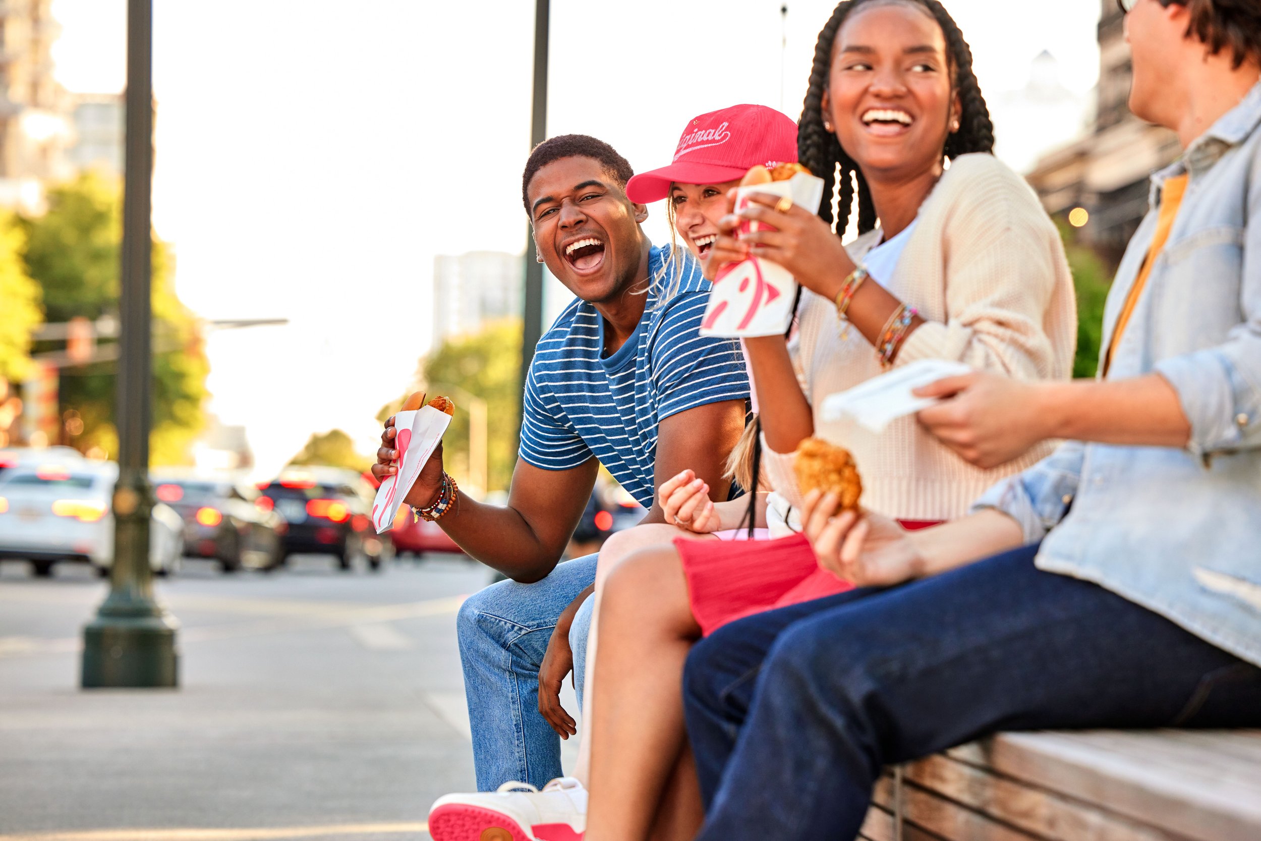 Four young friends sitting on a park bench, eating fast food, and laughing together outdoors.