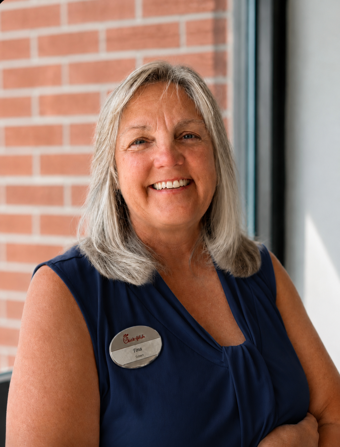 Smiling woman with gray hair, wearing a navy blue sleeveless top and a Chick-fil-A name tag, standing near a brick wall and window.