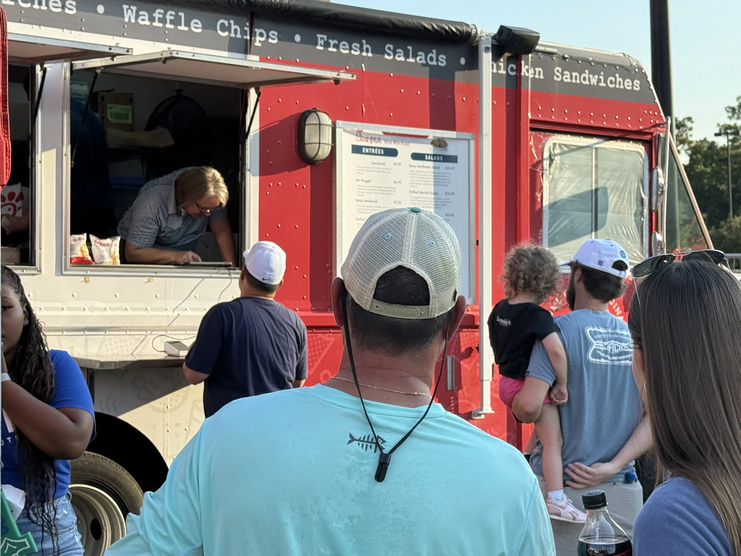 People waiting in line at a food truck, with a woman preparing food inside the truck and a man in a white cap facing toward the truck. A man wearing a baseball cap is holding a child, and another woman with sunglasses is visible nearby.