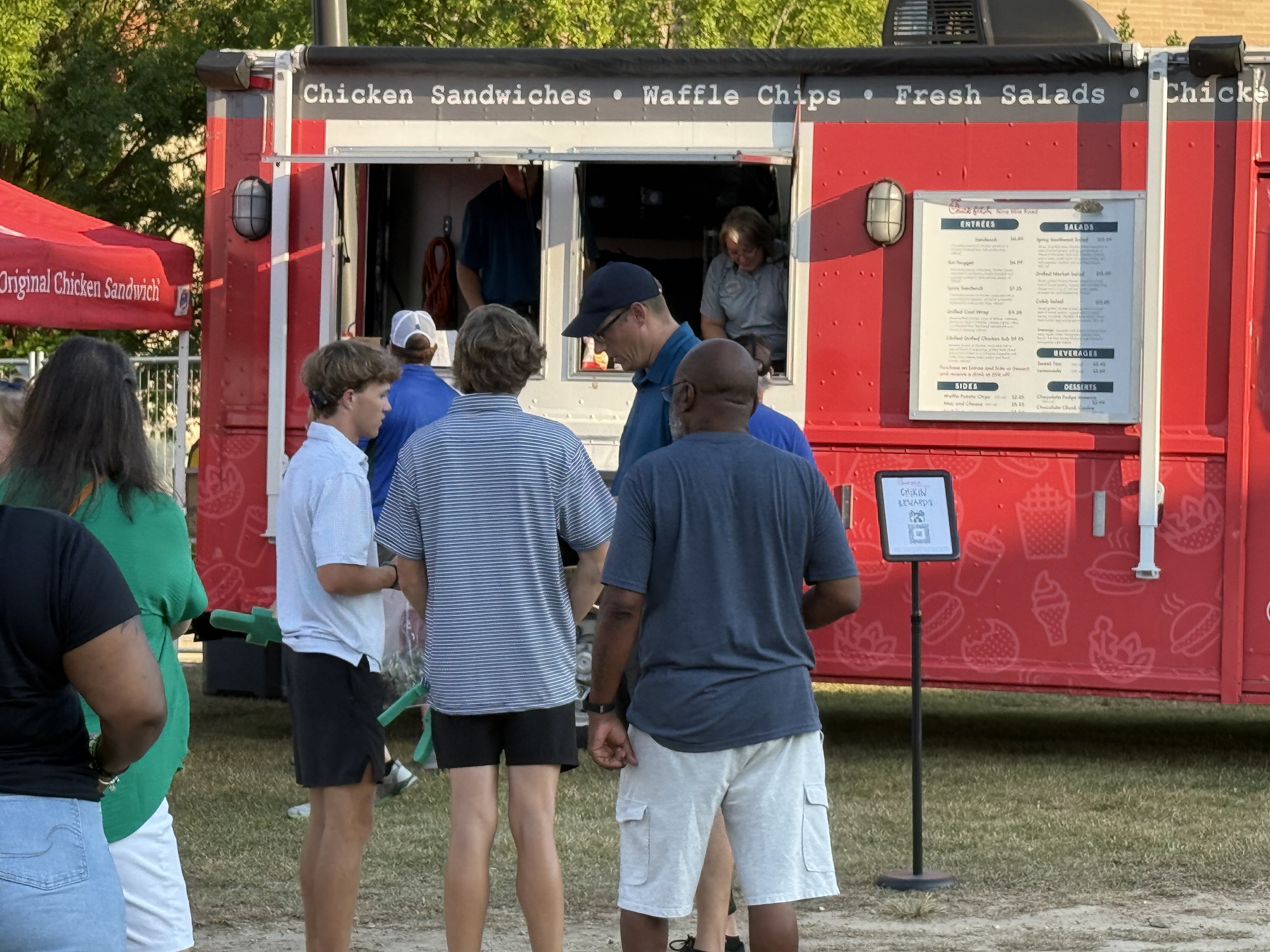 People standing in line at a red food truck that sells chicken sandwiches, waffle chips, and fresh salads.