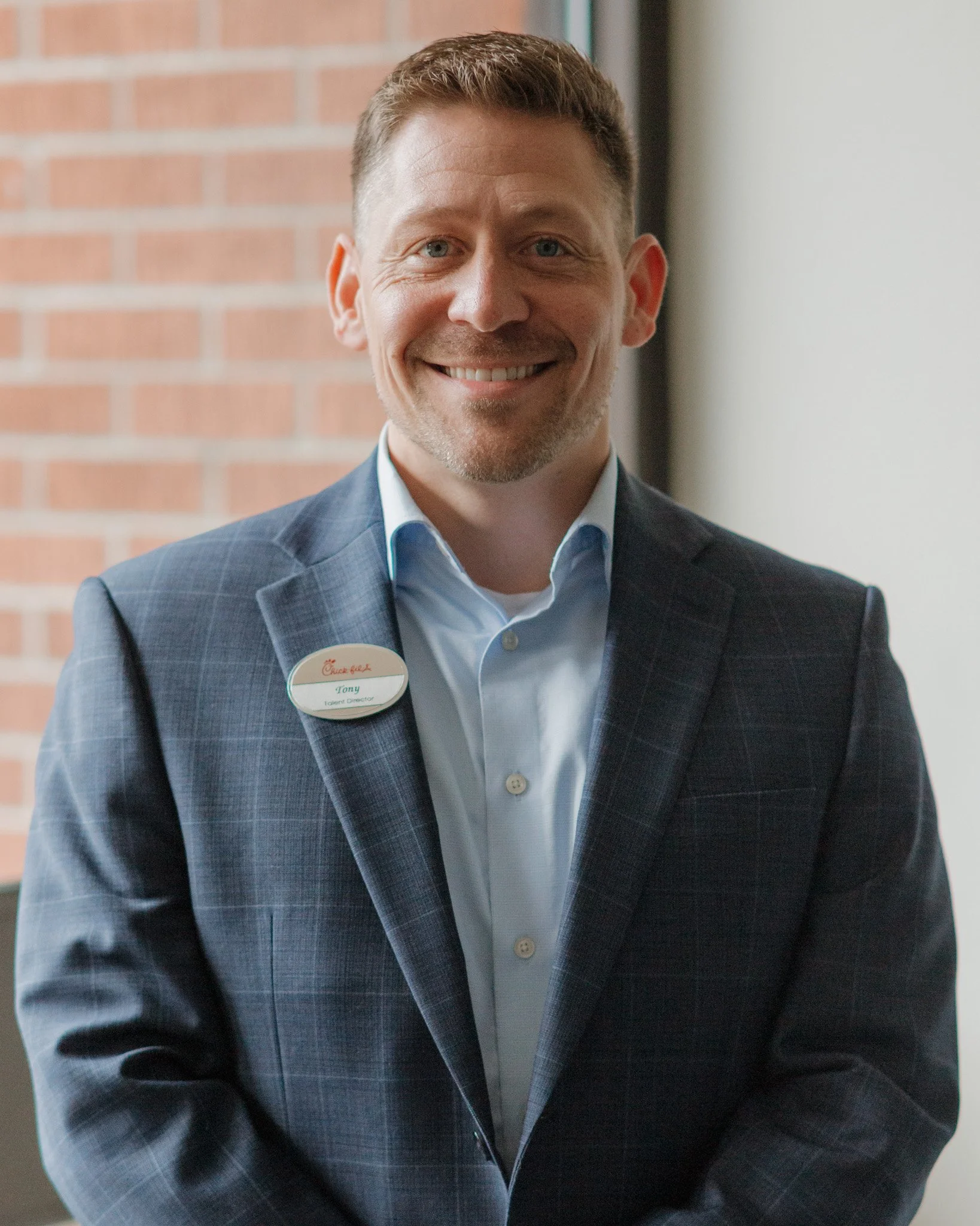 A smiling man with short brown hair and blue eyes, wearing a dark plaid suit jacket and a light blue collared shirt, standing in front of a brick wall with a white wall to the right. He is wearing a name tag and appears to be at a professional or corporate event.