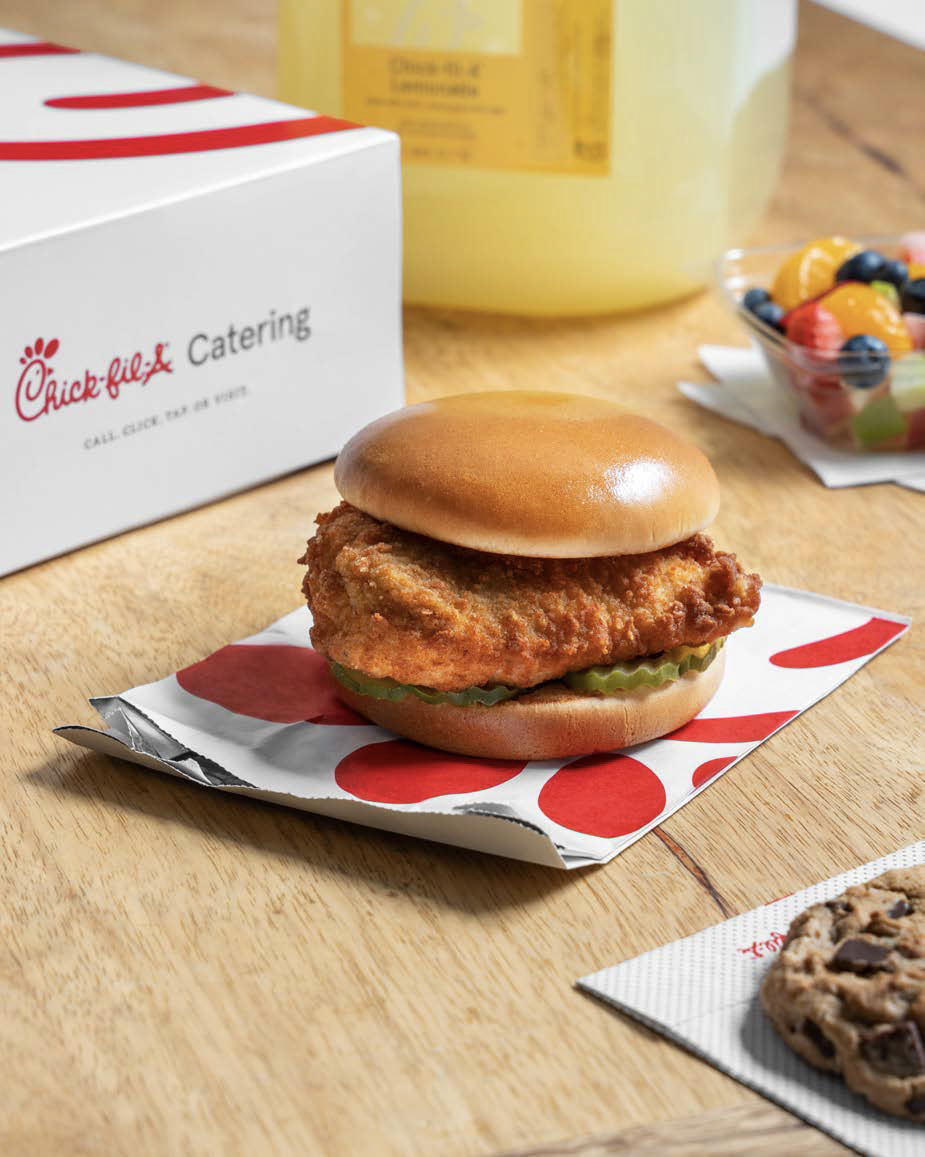A classic Chick-fil-A chicken sandwich on a wooden table with a catering box, lemonade, fruit cup, and a chocolate chip cookie in the background.