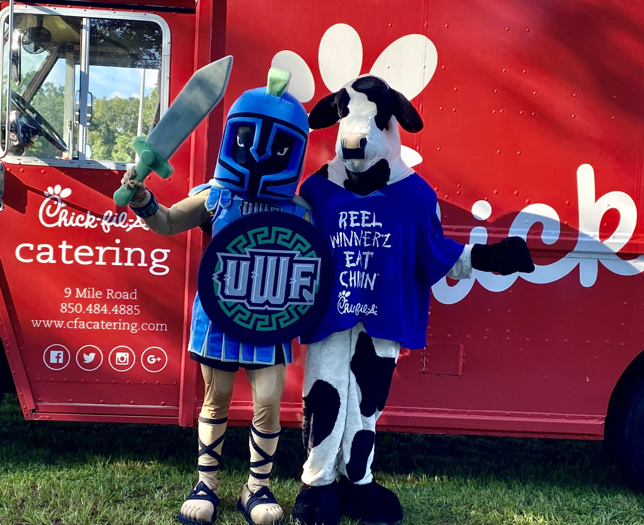 Person dressed as a warrior holding a sword, next to a person in a cow costume, standing in front of a Chick-fil-A food truck.