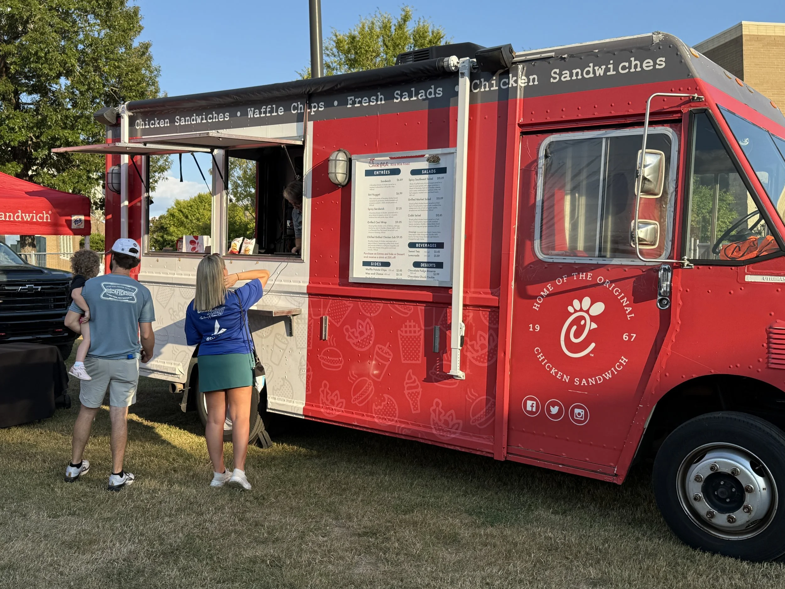 A red Chick-fil-A food truck with a menu and food options on the side, serving chicken sandwiches, waffle chips, and salads. Customers are ordering at the window, with a woman and child waiting in front.
