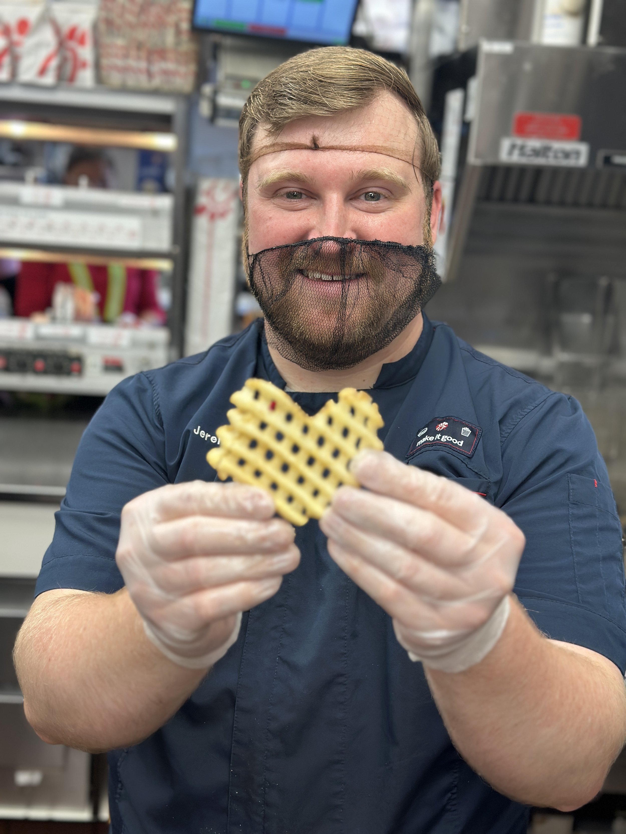 A Chick-fil-A Nine Mile Road team member in the kitchen smiling and holding up a heart-shaped waffle fry.
