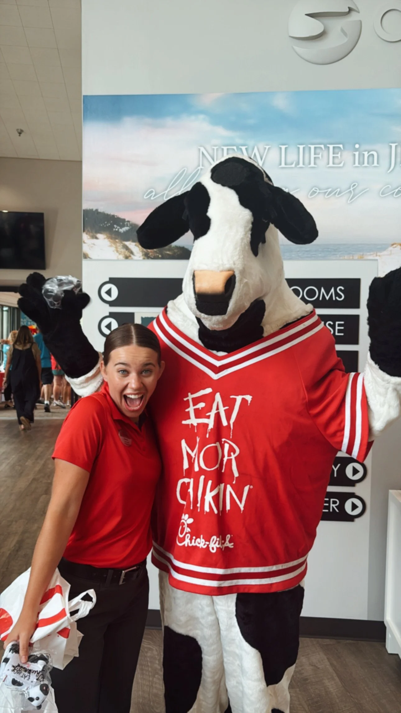 A woman in a red Chick-fil-A uniform smiling next to a person dressed as a cow mascot wearing a red cow costume with the phrase "EAT MOR CHIKIN" written on it. The woman has her arm around the cow mascot and appears happy.