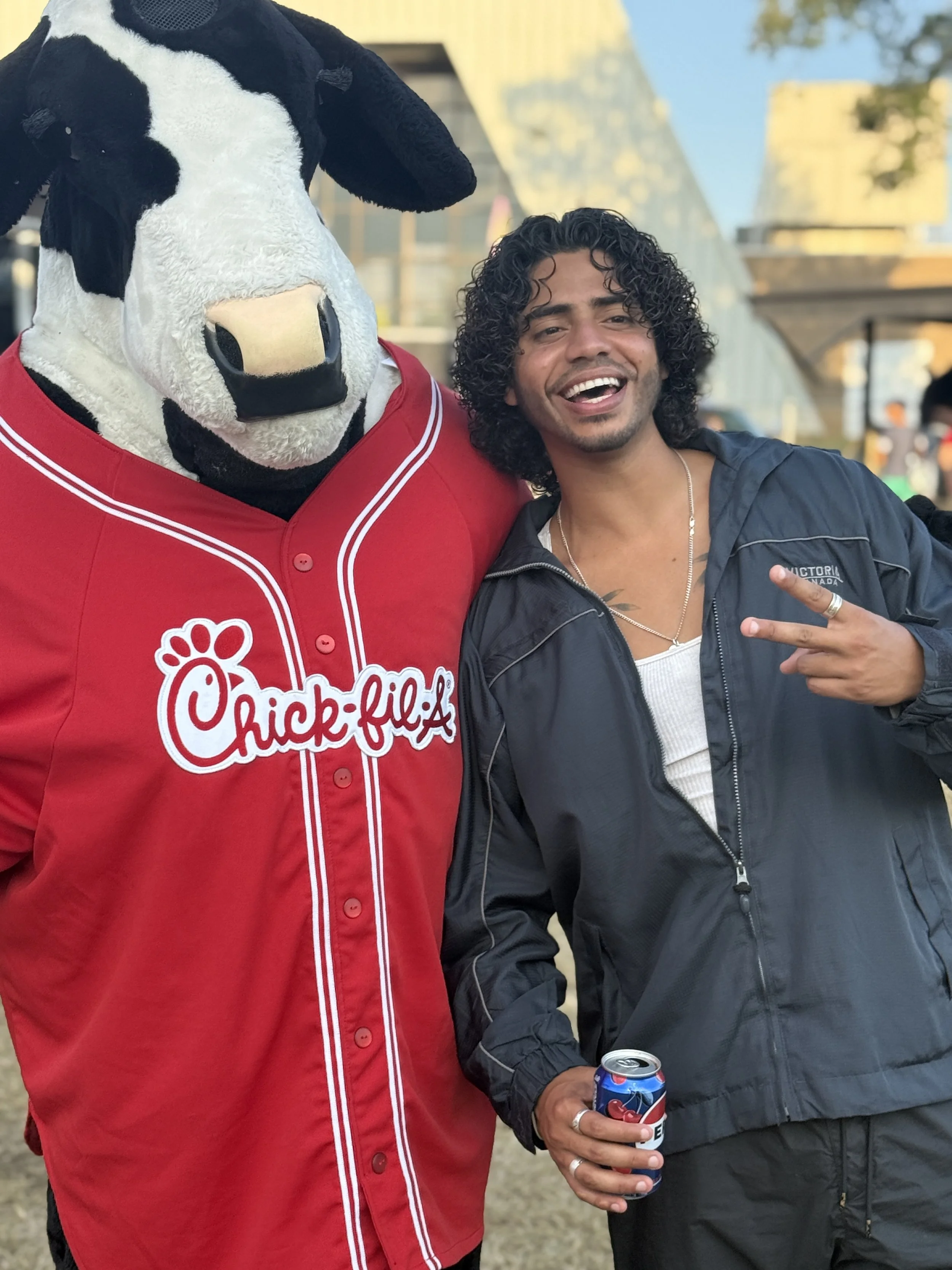 A man with curly hair smiling and making a peace sign gesture standing next to a person in a Chick-fil-A cow mascot costume, outdoors during daytime.