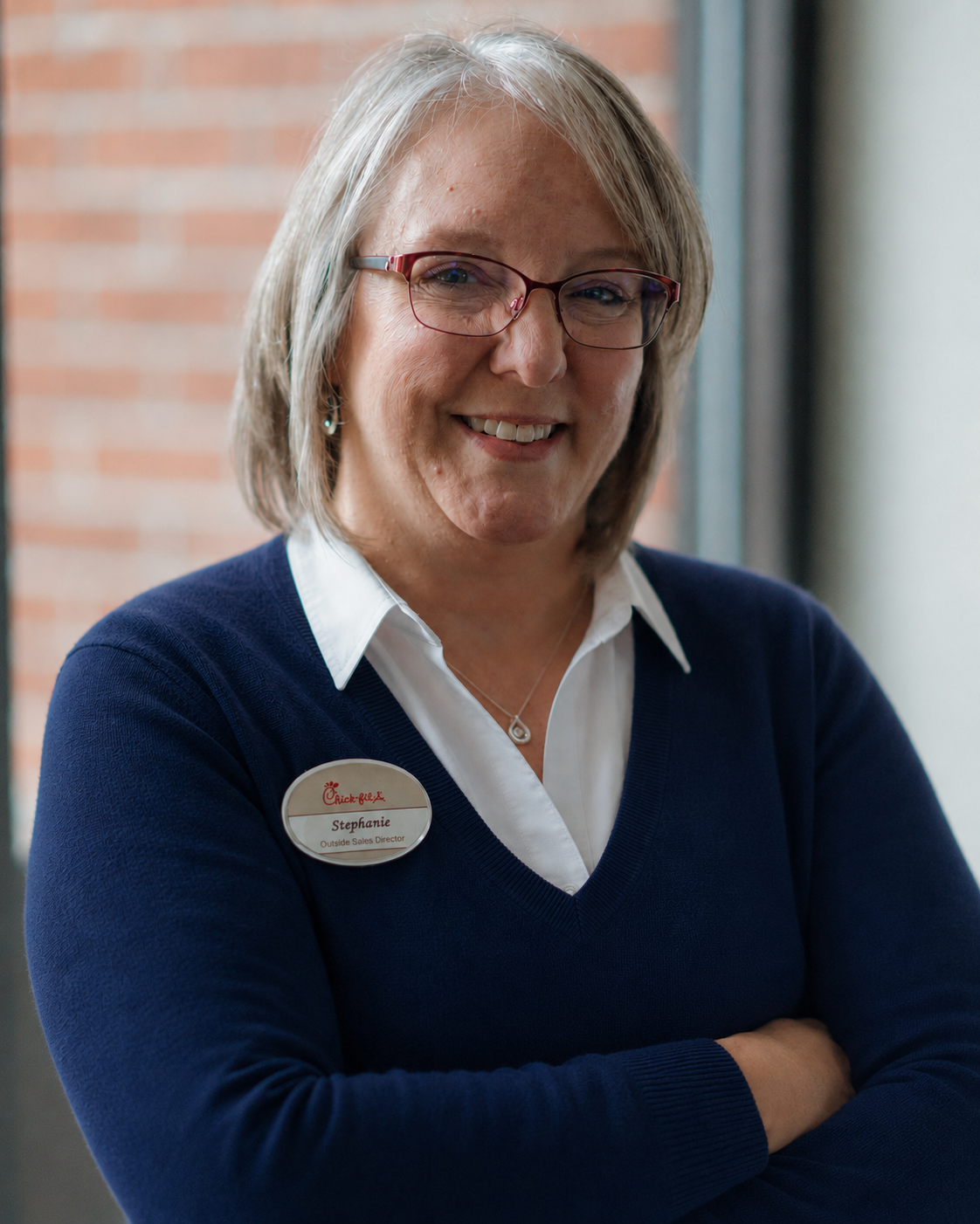 A woman with glasses and gray hair smiling, wearing a white shirt and a dark blue sweater with a Chick-fil-A name tag reading "Stephanie, Outside Sales Director," standing near a brick wall and window.