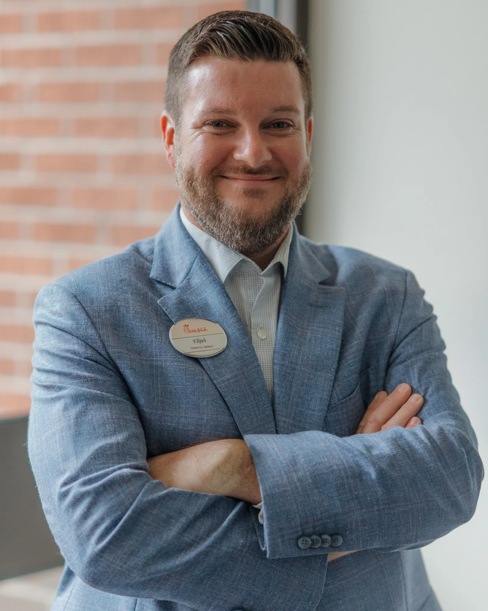 A man with a neatly styled hairstyle and a beard, wearing a suit jacket and a name badge, standing with arms crossed in front of a brick wall.
