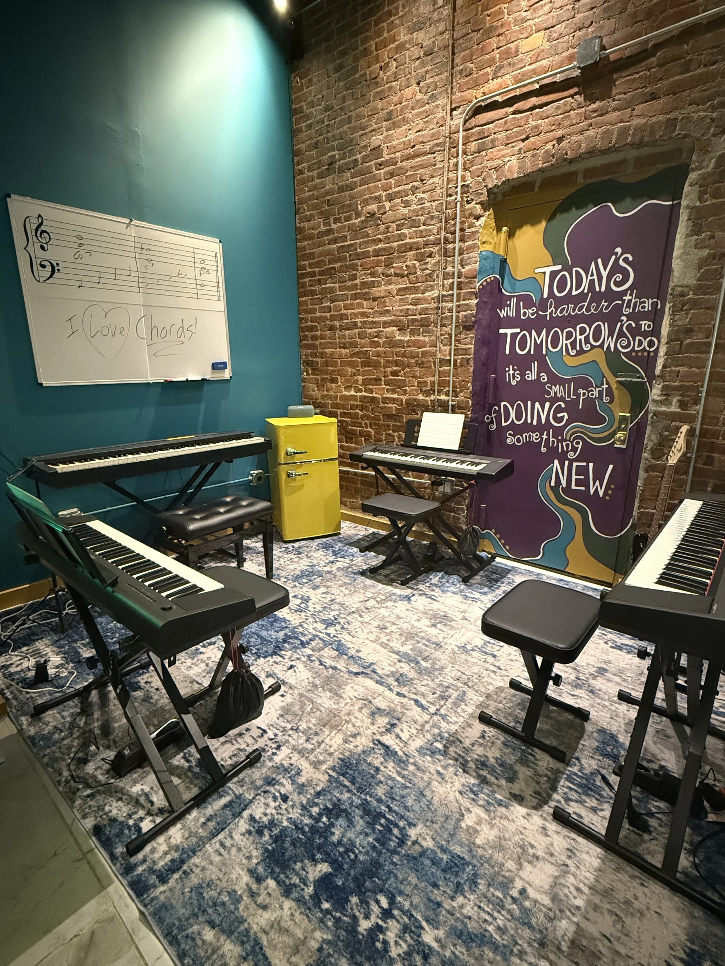 Music practice room with electronic pianos, a whiteboard with musical notes and the phrase 'I love chords!', a yellow refrigerator, a colorful mural with motivational quote, and a patterned blue and beige rug.